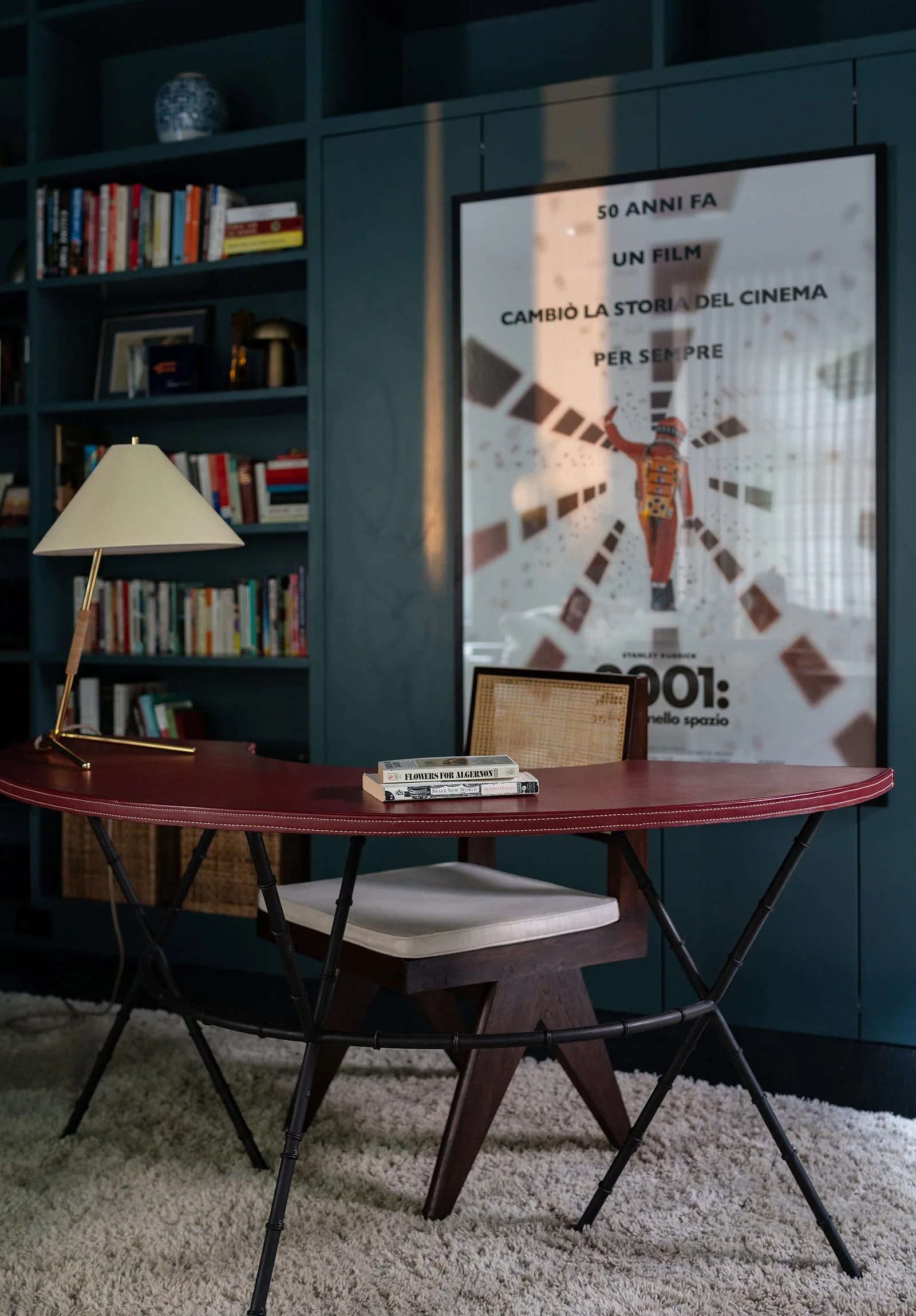 A curved red desk and a cane-back chair sit in front of deep teal built-in bookshelves and a large framed poster.