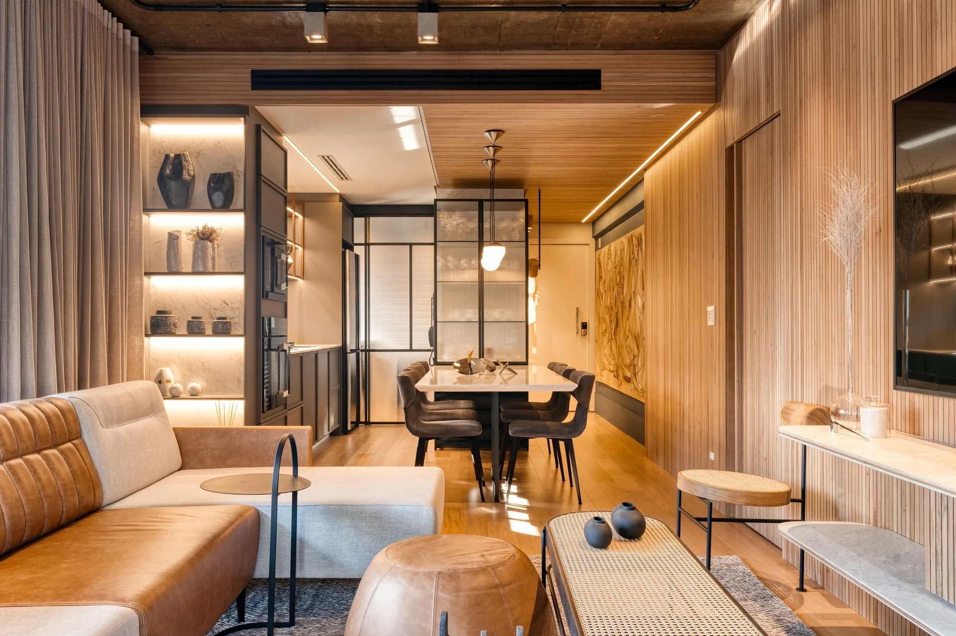 A modern open-concept living and dining area features a tan leather sofa, vertical wood slat wall paneling, and a long marble dining table.