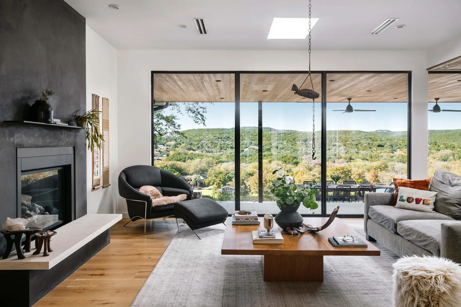 A modern living room featuring a dark fireplace wall, gray sofa, low wooden coffee table, and large sliding glass doors showing a hillside view.