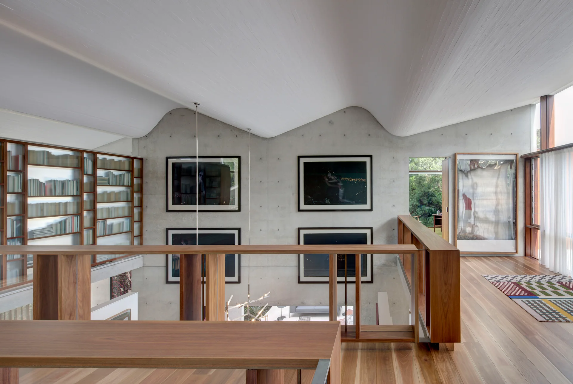 A modern residential mezzanine features a wavy white sculptural ceiling, board-formed concrete walls with framed art, and warm wooden floors and railings.