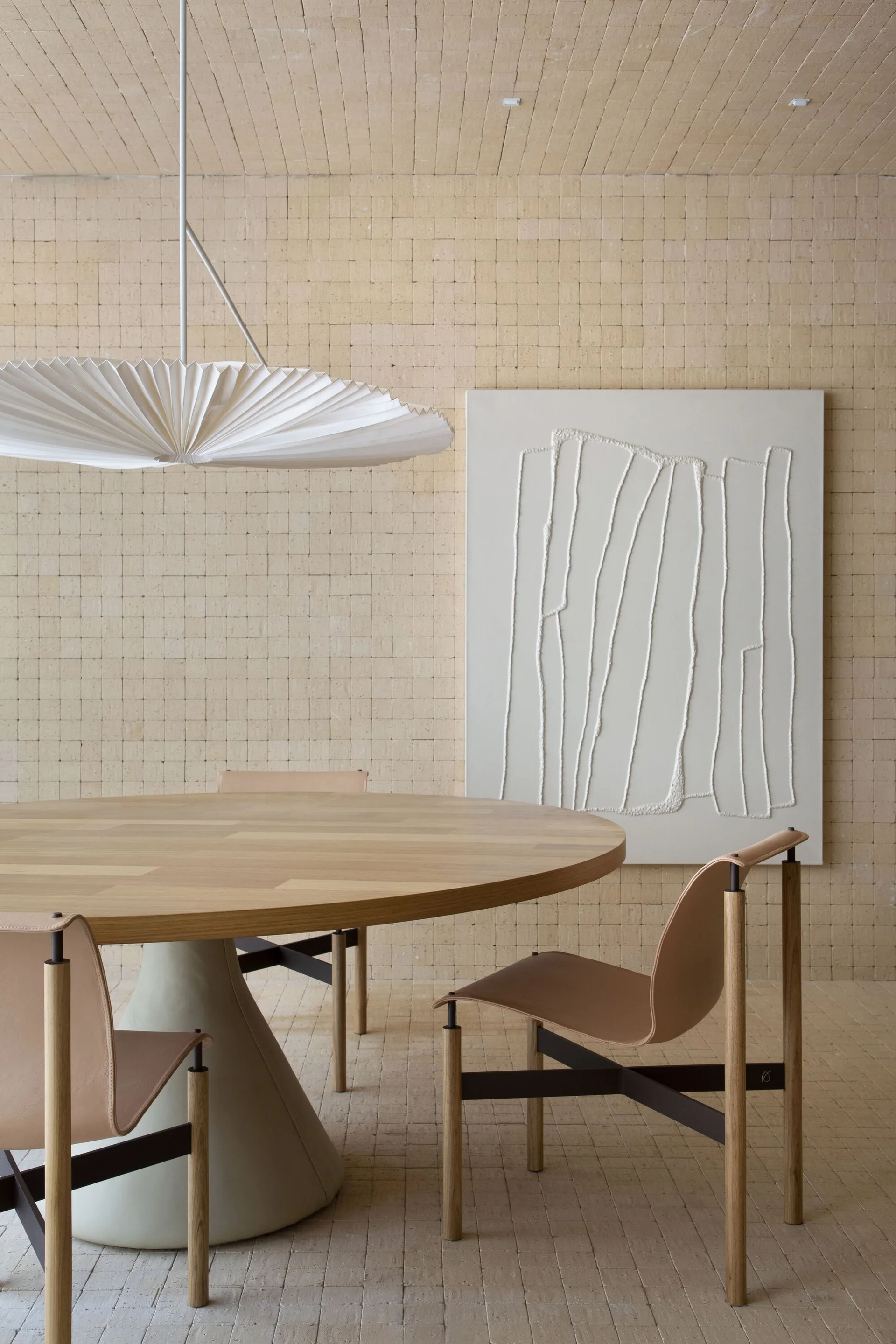 A round wood dining table and leather chairs sit in a room with beige tiled walls and ceiling, under a pleated circular pendant light.