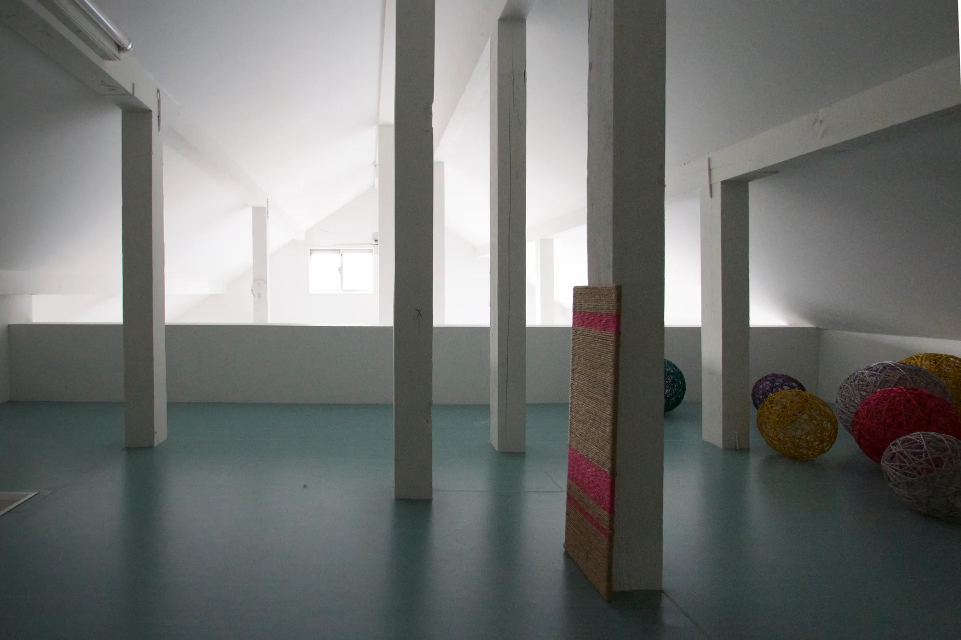 A minimalist white attic room featuring structural support columns, a muted teal floor, and several colorful yarn spheres.