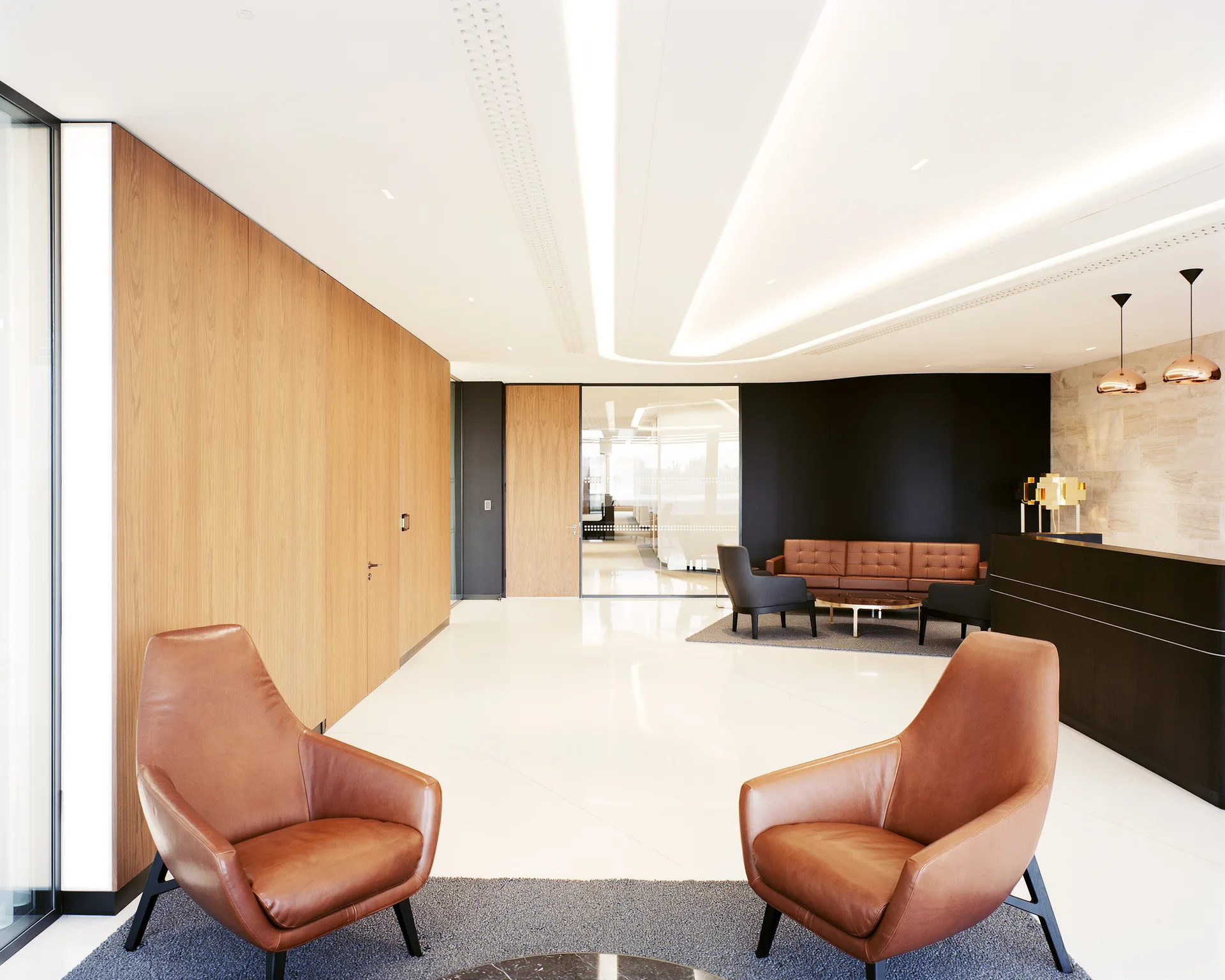 A minimalist office lobby featuring wood-paneled walls, tan leather armchairs, a dark reception desk, and curved ceiling lights.