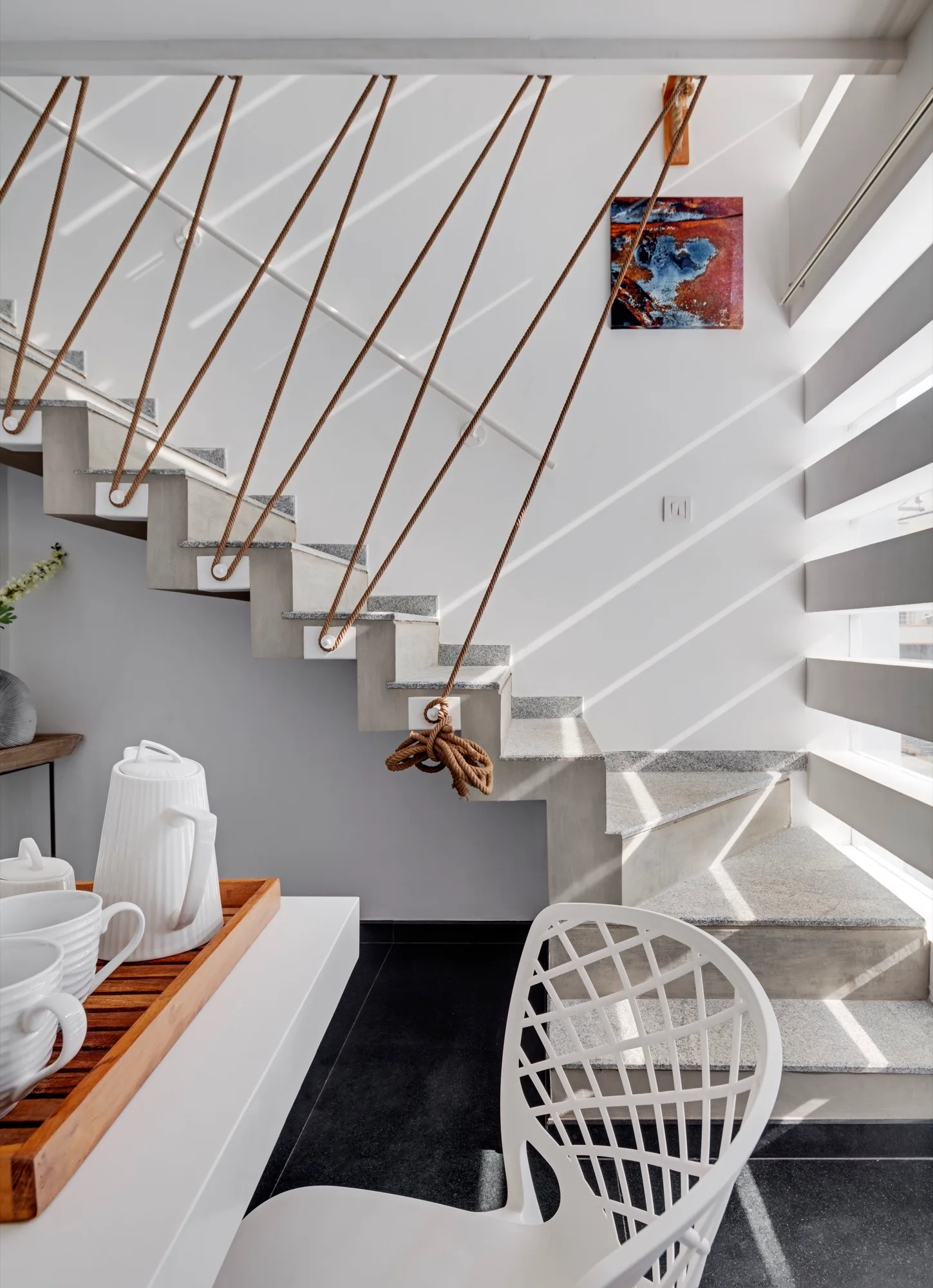 A modern floating stone staircase features thick brown ropes used as a railing next to a slatted window casting geometric shadows.