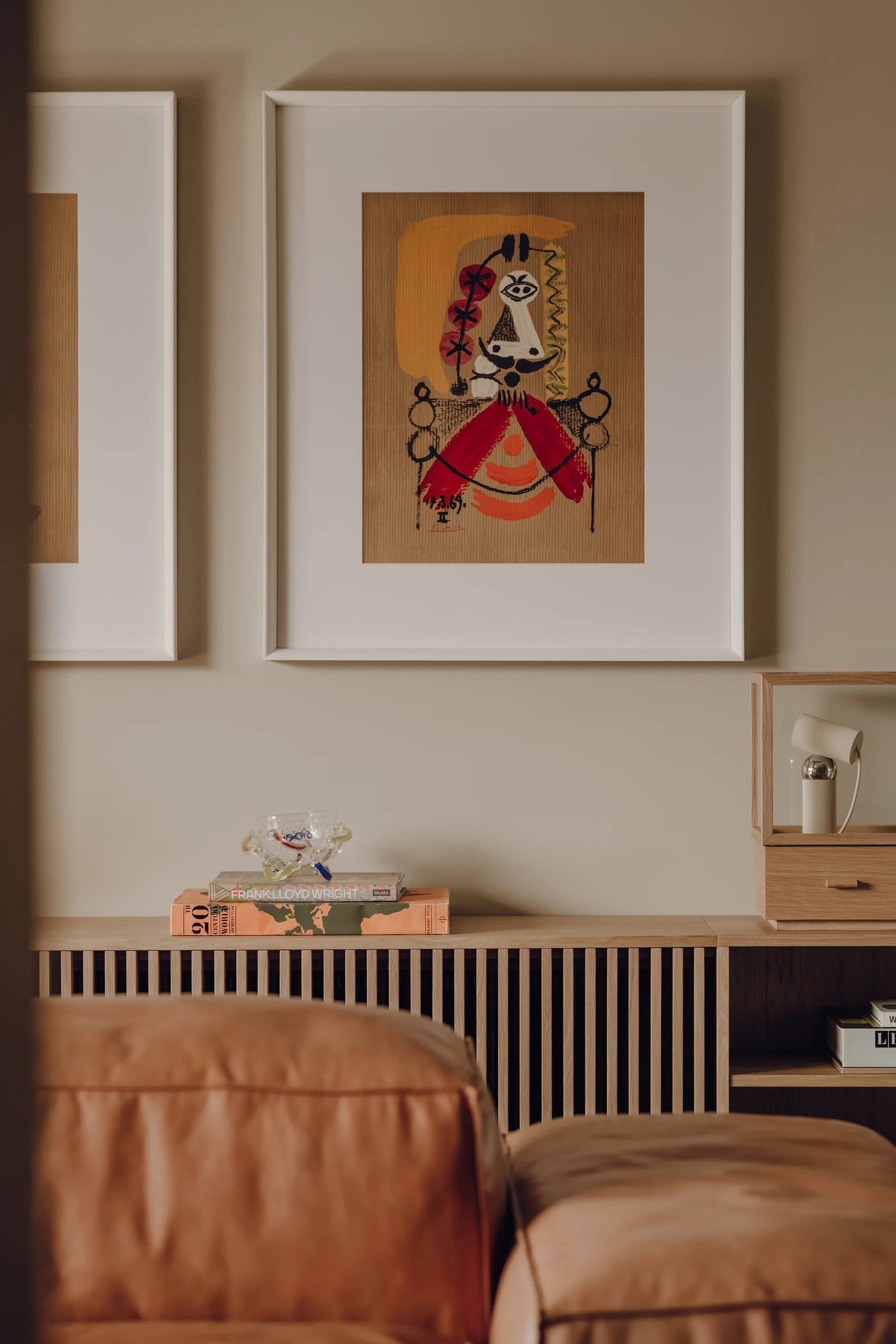 A light wood slatted console table holds books and art beneath two large framed prints on a beige wall, with a tan leather sofa in the foreground.