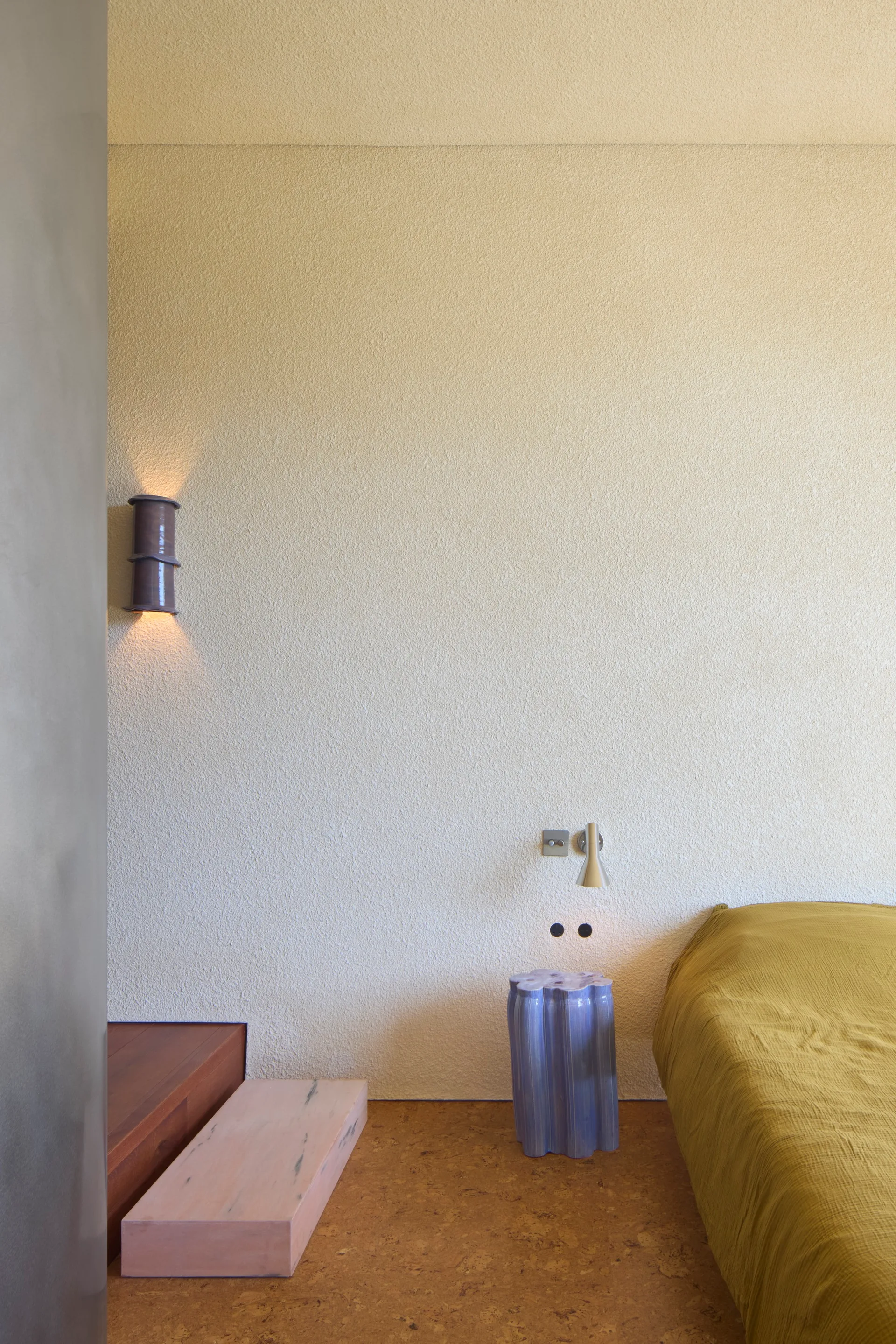 A modern bedroom featuring textured plaster walls, warm cork flooring, a mustard yellow bed, and a sculptural purple ceramic side table.