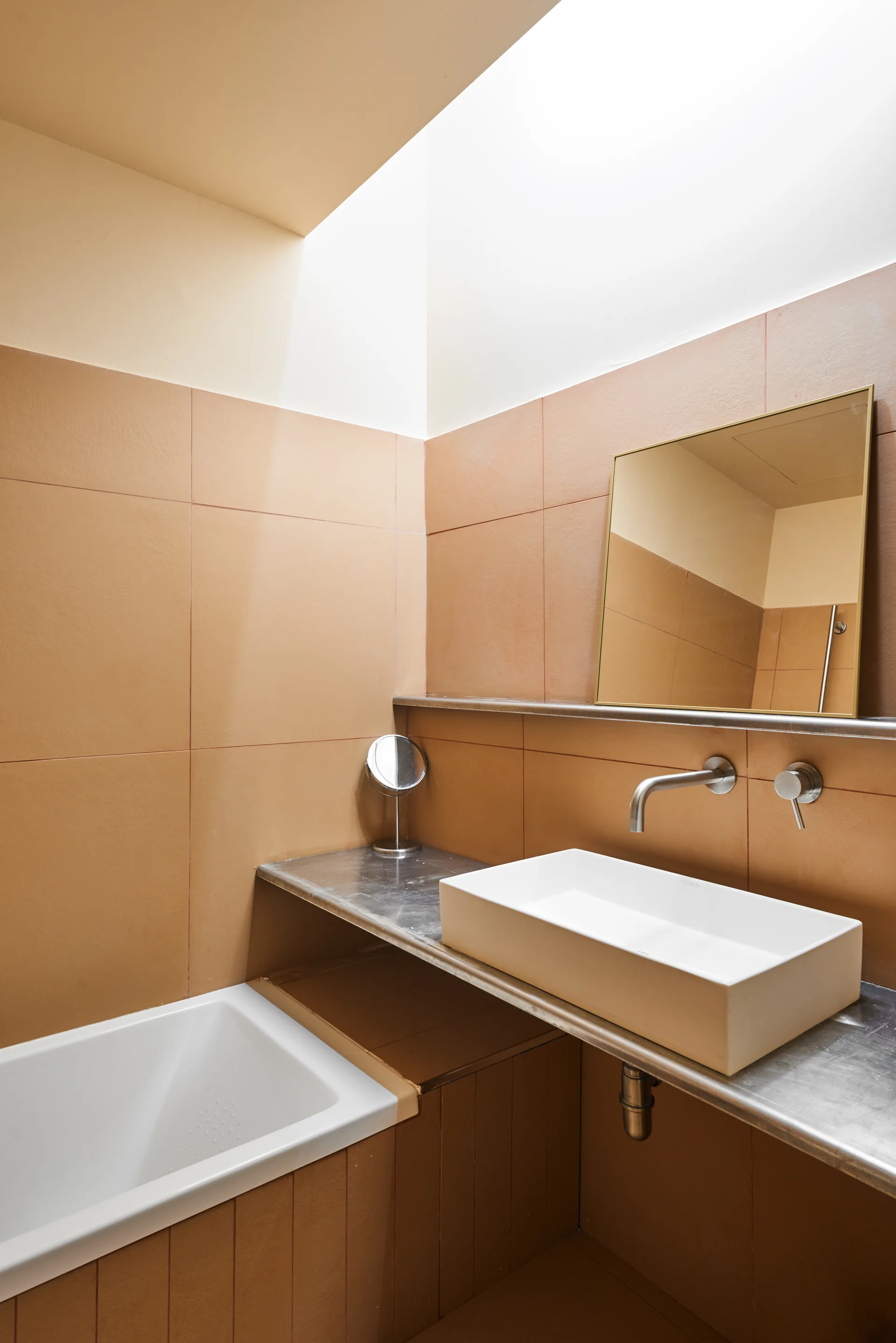 A white vessel sink sits on a brushed metal countertop against a tan tiled wall in a minimalist bathroom with a skylight.