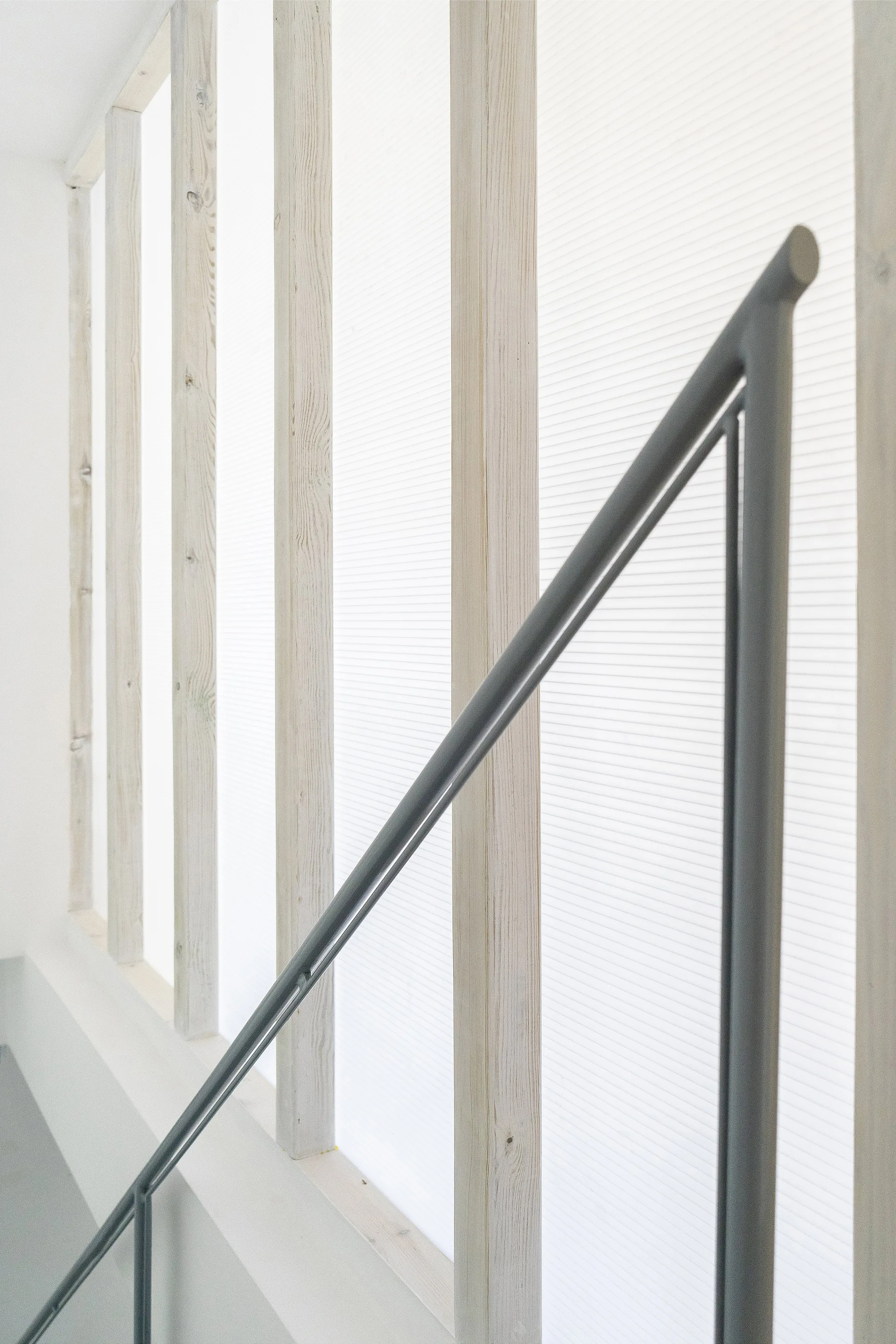 A minimalist dark grey metal stair railing set against vertical pale wood studs and a translucent white ribbed backing panel.