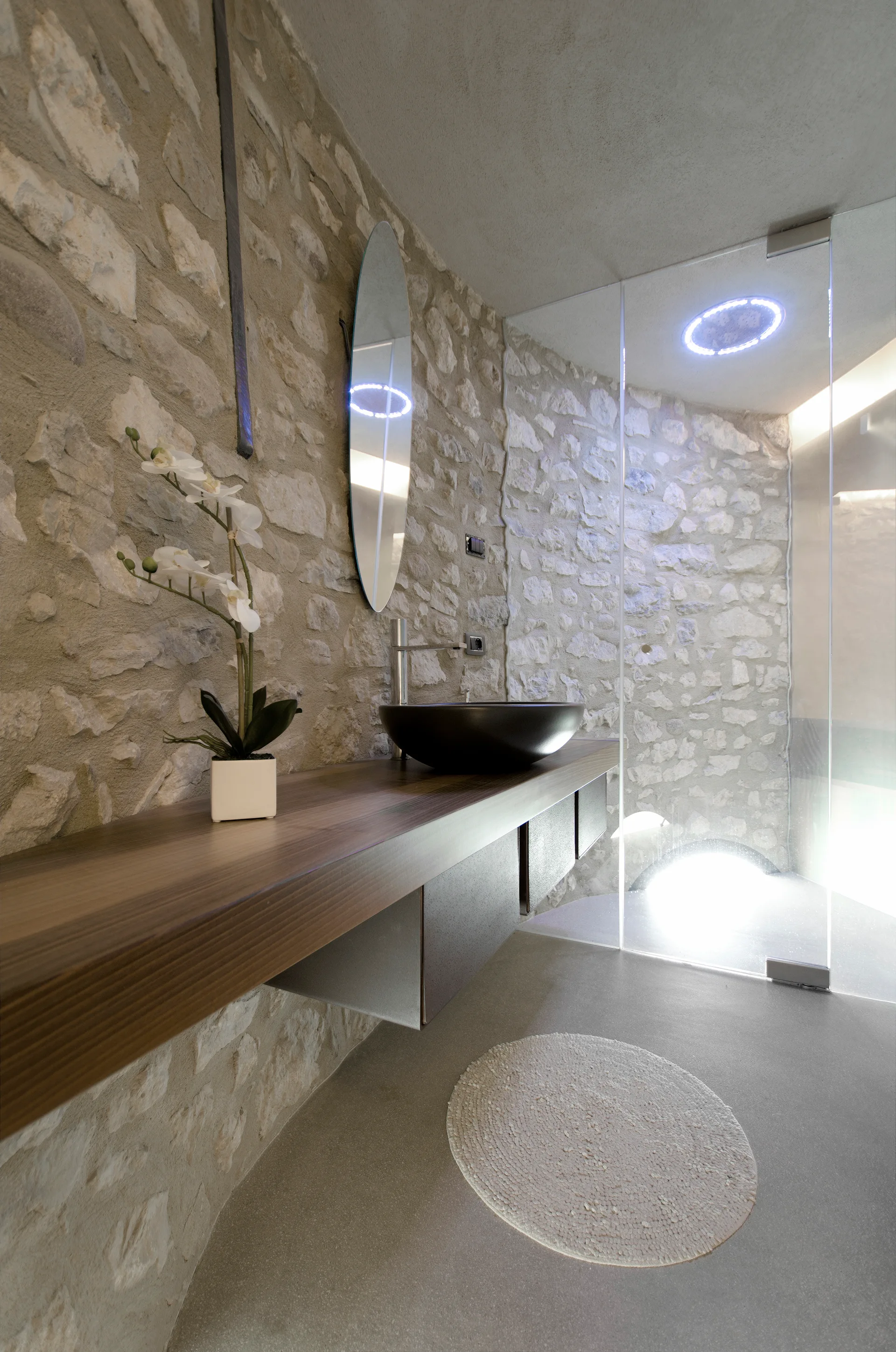 A modern rustic bathroom features a floating wood vanity with a black vessel sink set against a rugged stone wall next to a glass shower.