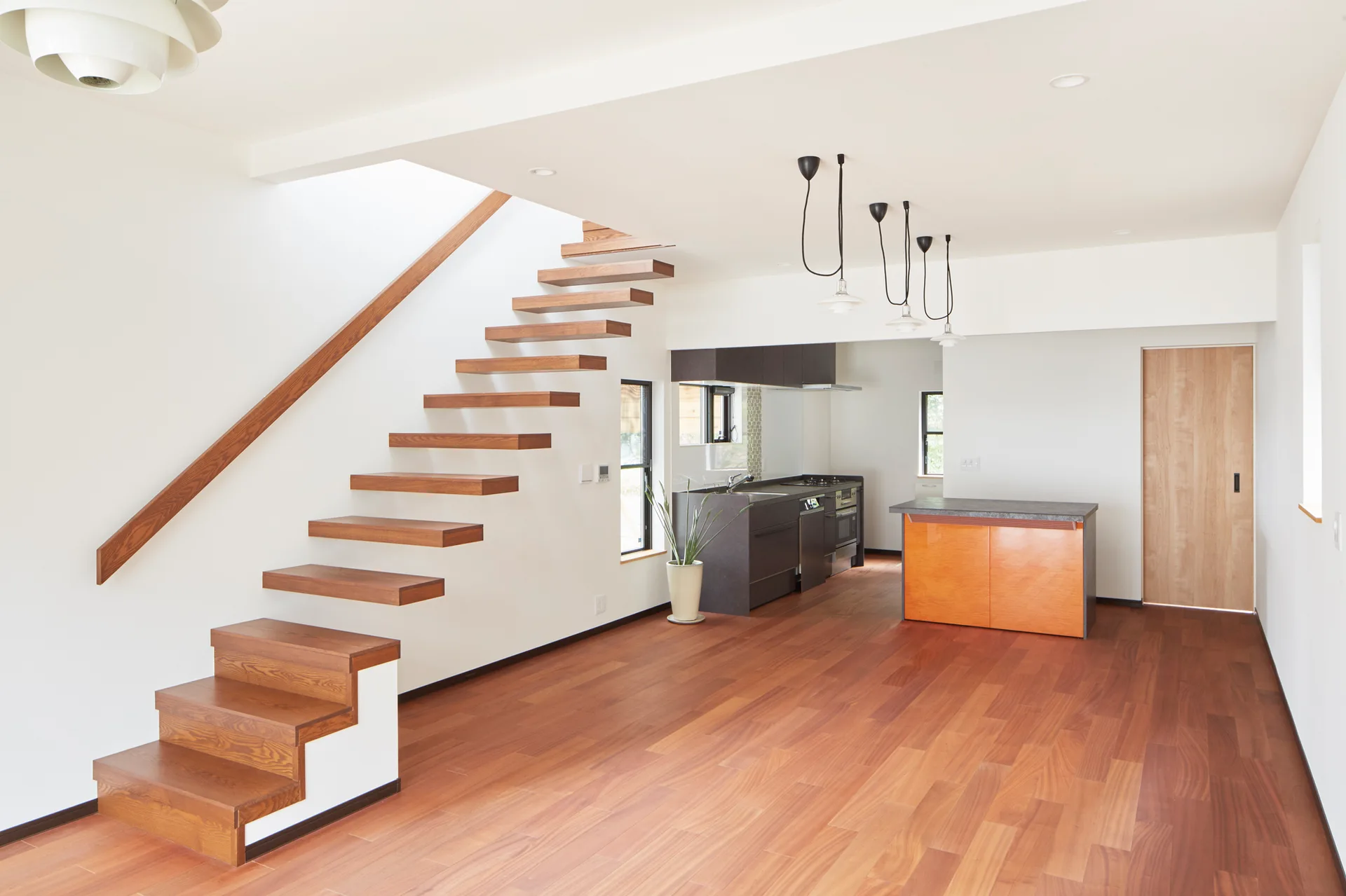 A floating wooden staircase rises against white walls in an open-plan home featuring warm hardwood floors and a minimalist kitchen in the background.