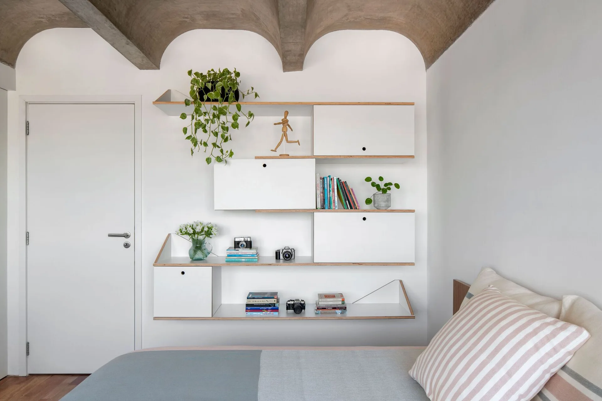 A modern bedroom features a white modular shelving unit with exposed wood edges mounted on a white wall beneath a vaulted concrete ceiling.