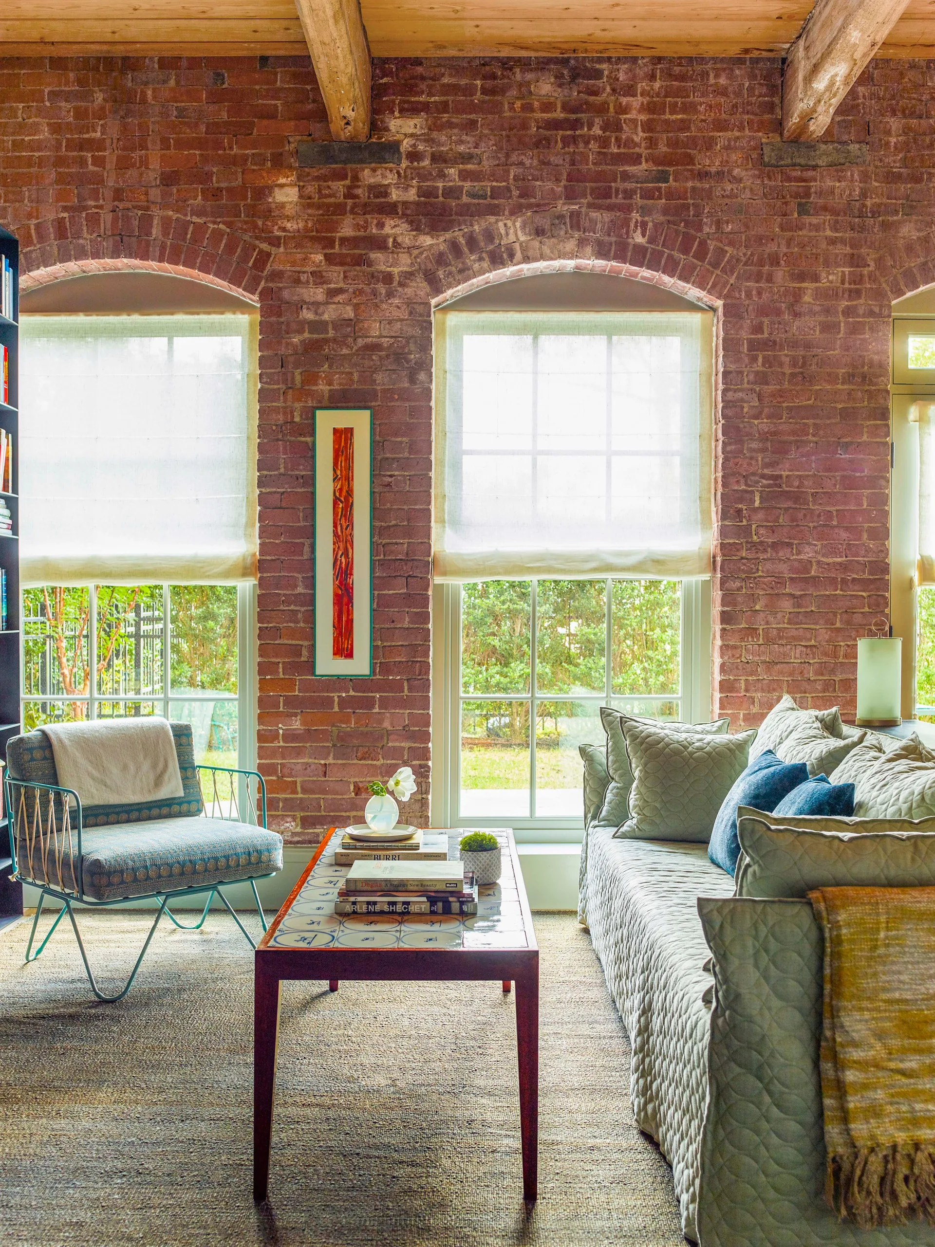 A living room featuring an exposed red brick wall with two arched windows, a green quilted sofa, and a tiled coffee table.