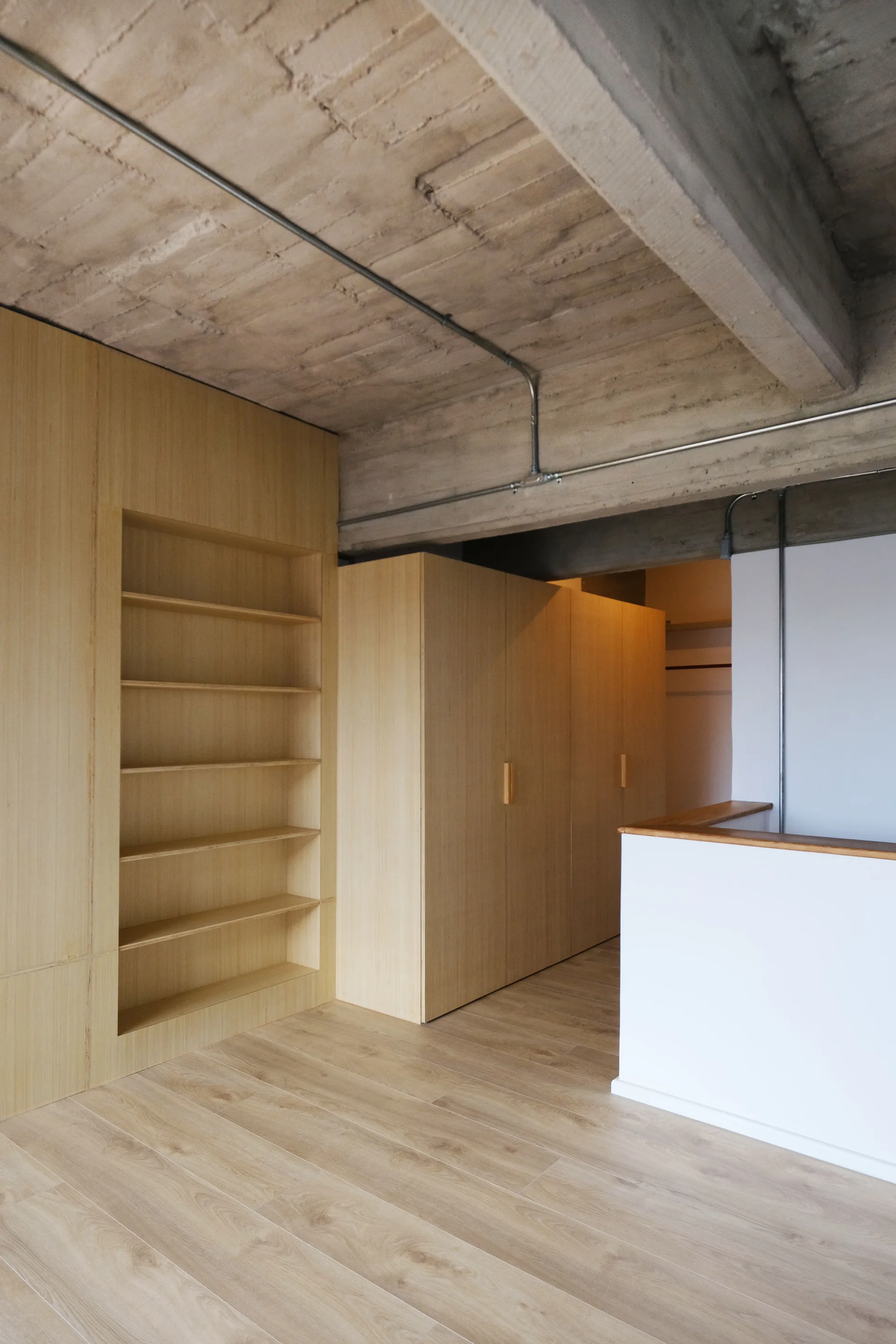 Light wood built-in wardrobes and recessed shelving stand beneath a raw concrete ceiling with exposed metal conduit and wood flooring.