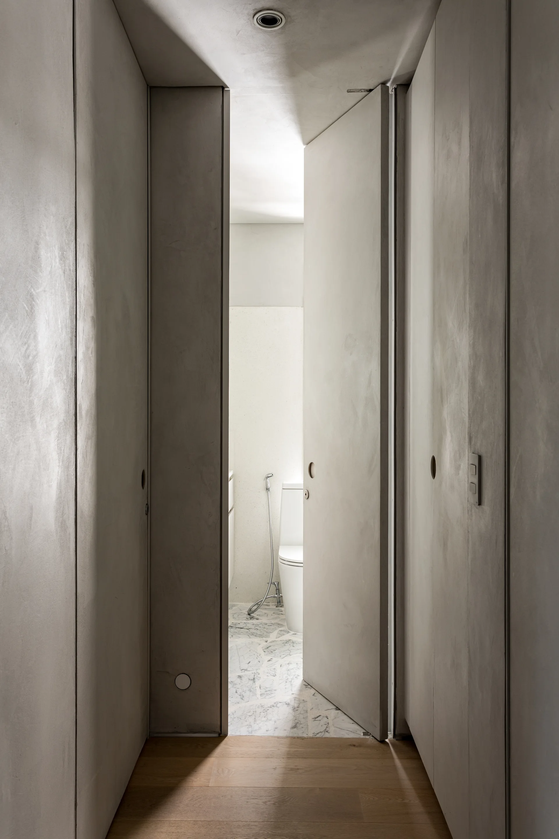 A tall grey frameless door with a round flush pull opens from a textured concrete-look hallway into a bright bathroom with veined marble tile flooring.