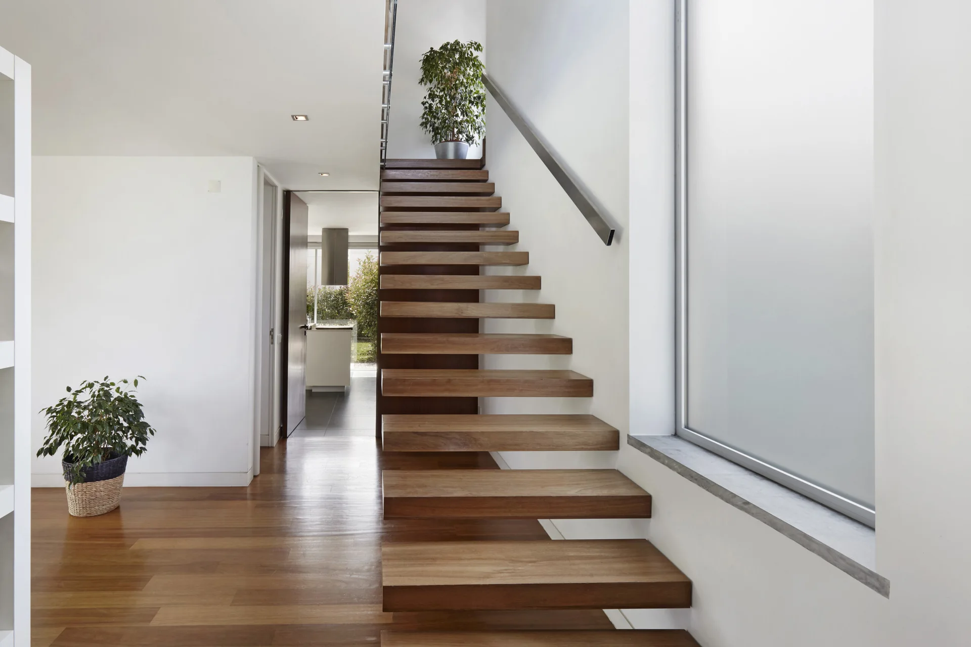 A wooden floating staircase stands next to a large window in a white-walled hallway with matching hardwood floors and potted plants.