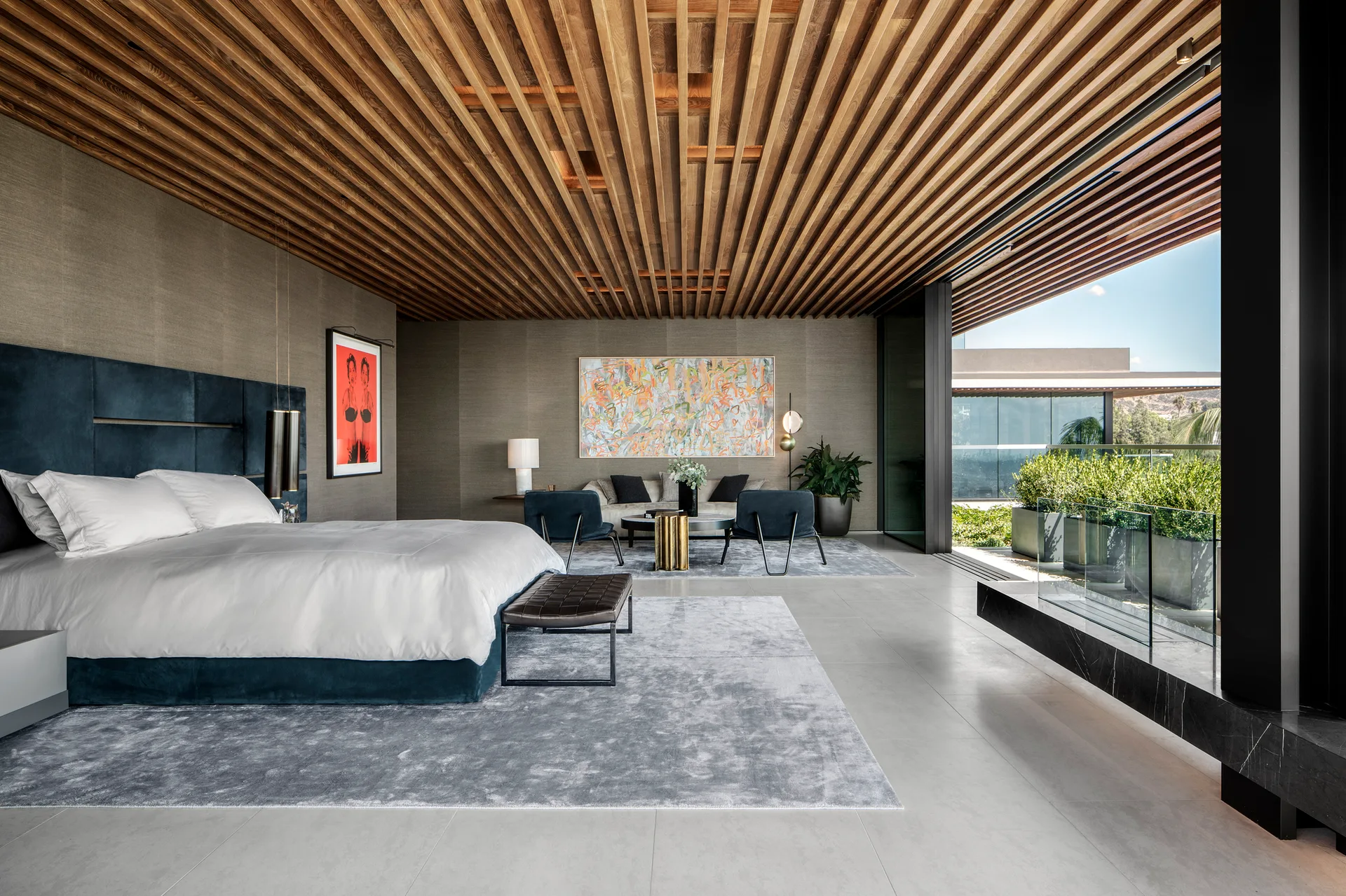 A spacious bedroom features a slatted wood ceiling, a blue upholstered bed, grey rugs, and sliding glass doors that open to a balcony.
