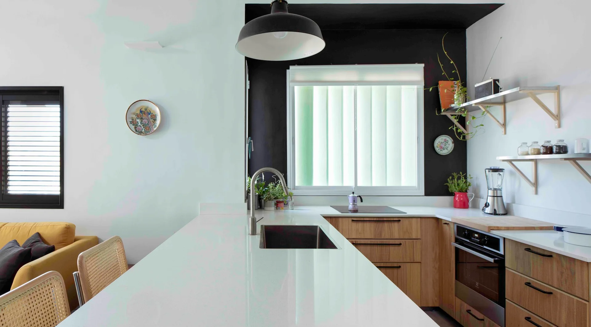 A modern kitchen featuring natural wood cabinets, a long white countertop island with a sink, and a black accent wall around a window.