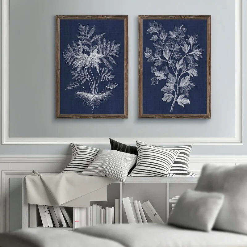 Two framed navy blue botanical prints hang on a light gray wall above a white console table with striped throw pillows and stacked books.
