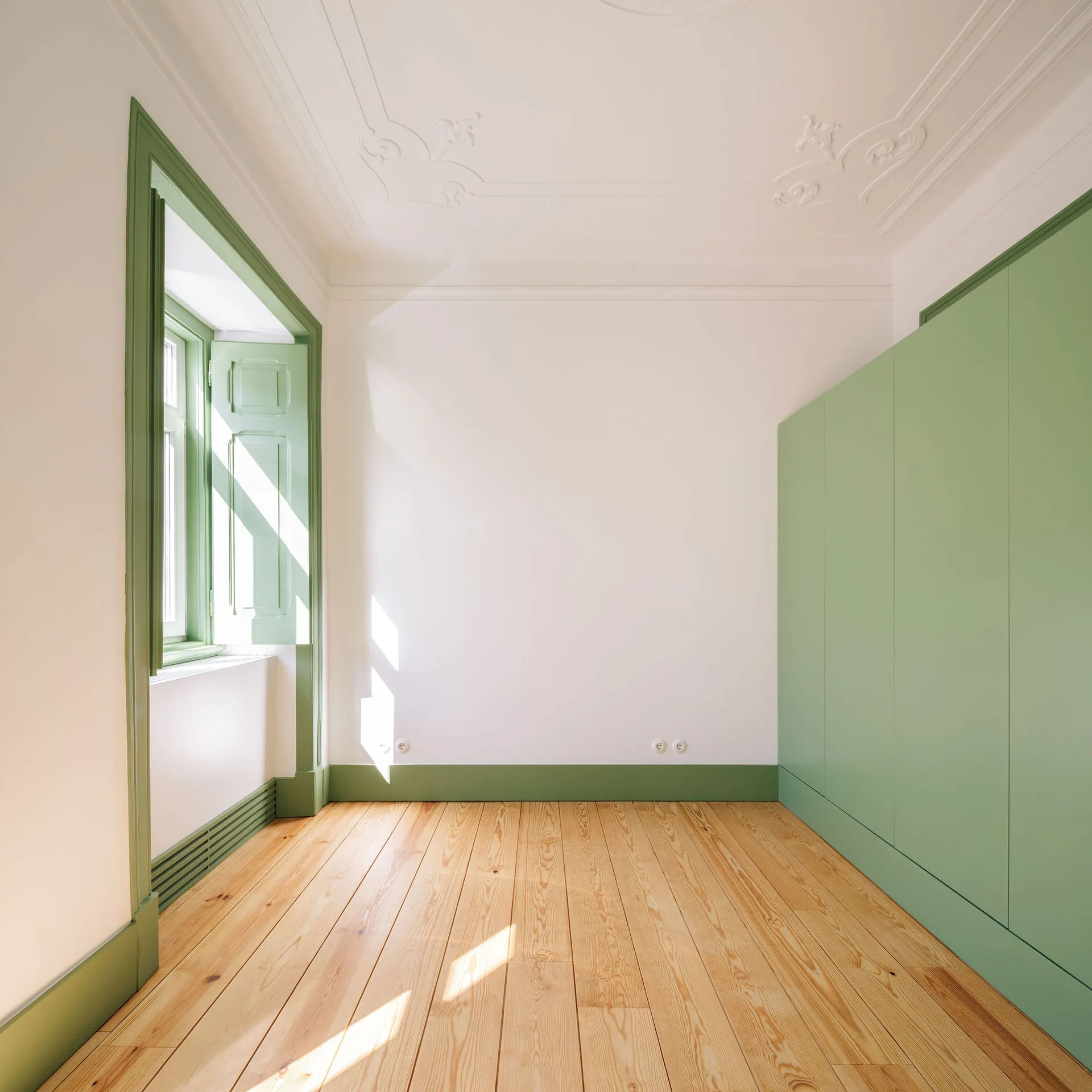An empty bedroom features light wood plank flooring, white walls with ornate ceiling molding, and sage green built-in closets and window trim.
