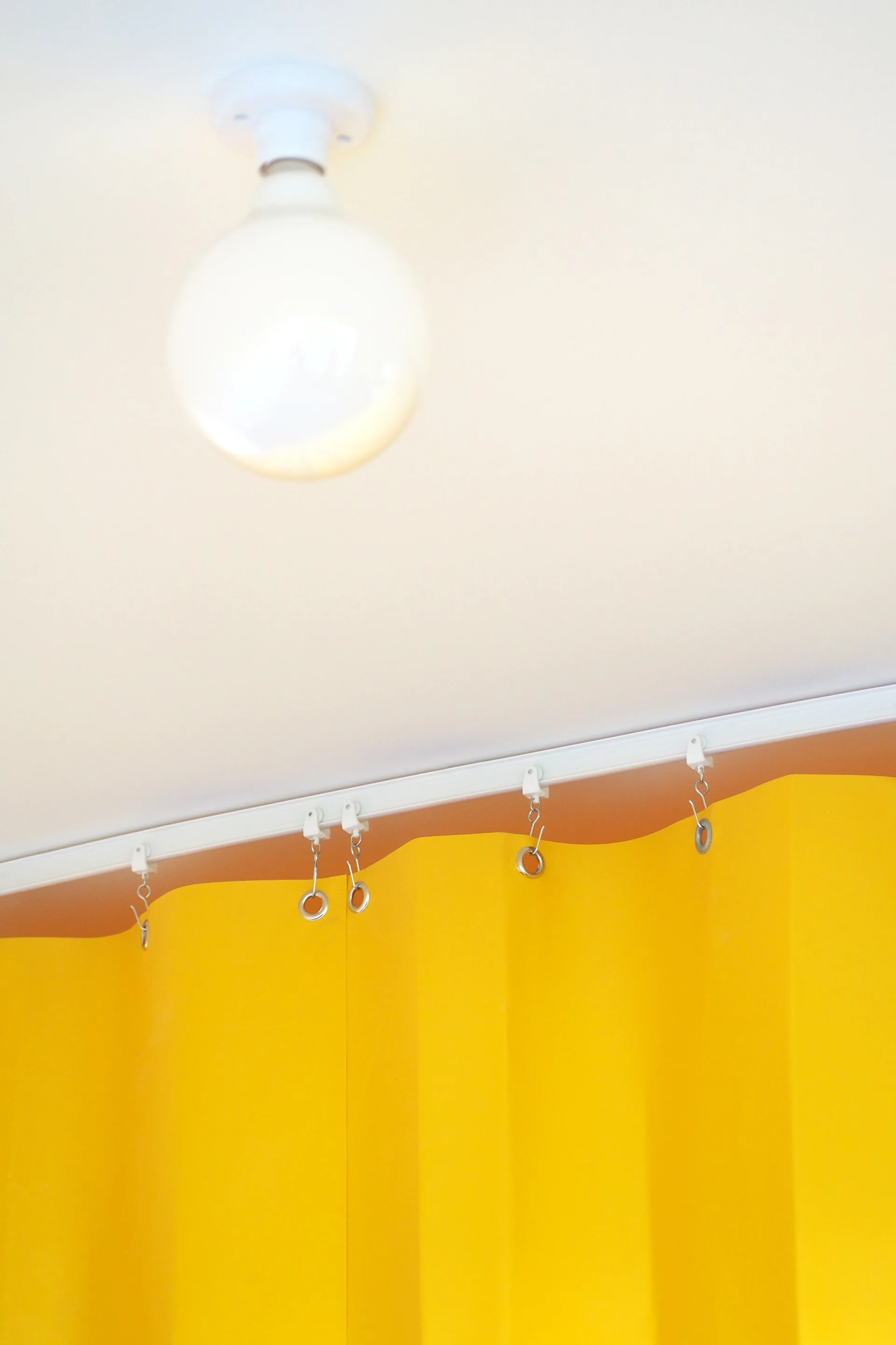 A bright yellow pleated curtain hangs from a low-profile white track mounted to a plain white ceiling near a round globe light fixture.
