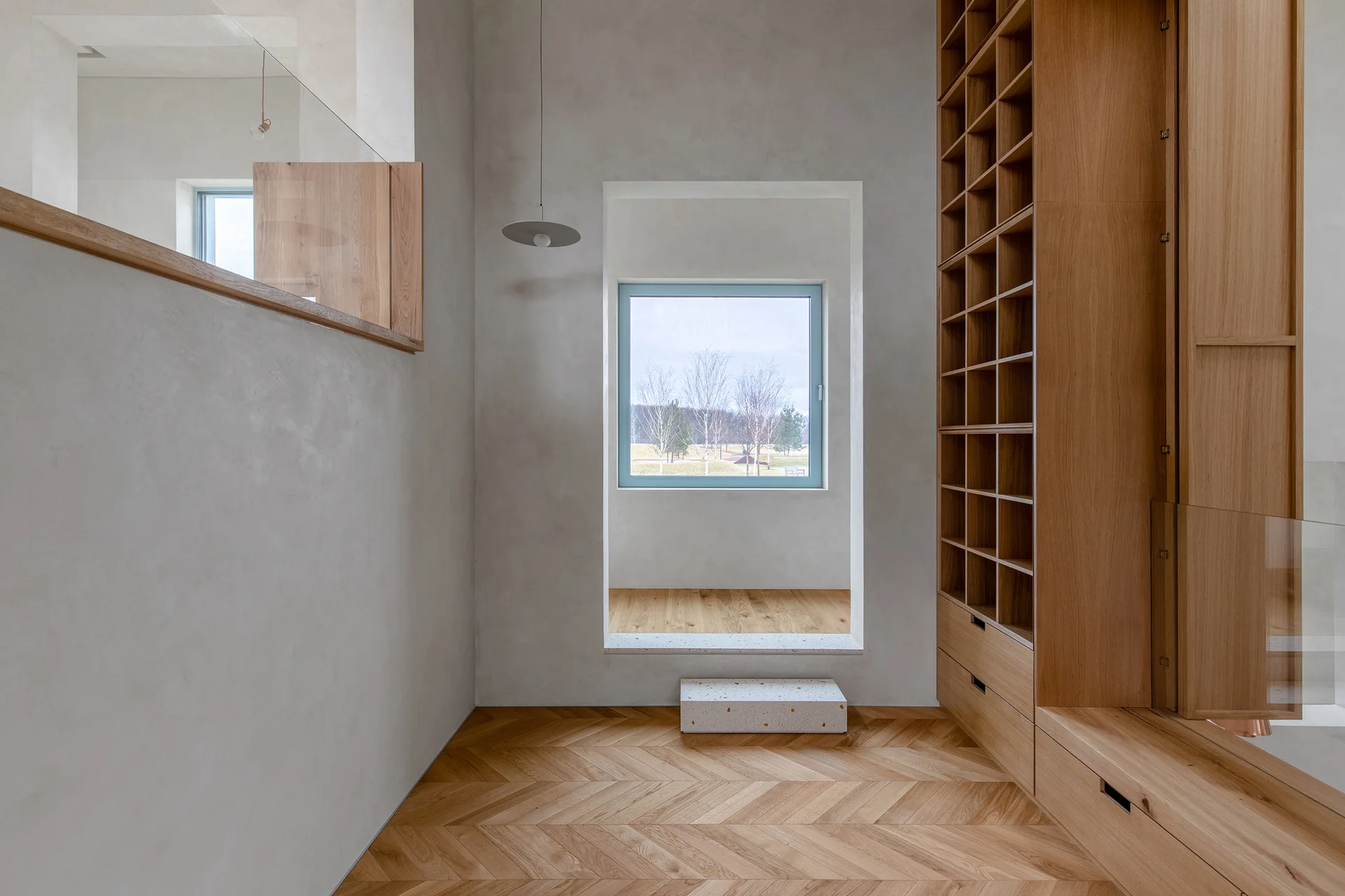 A minimalist hallway features light grey plaster walls, a custom oak grid storage wall, herringbone wood flooring, and a deep window niche.