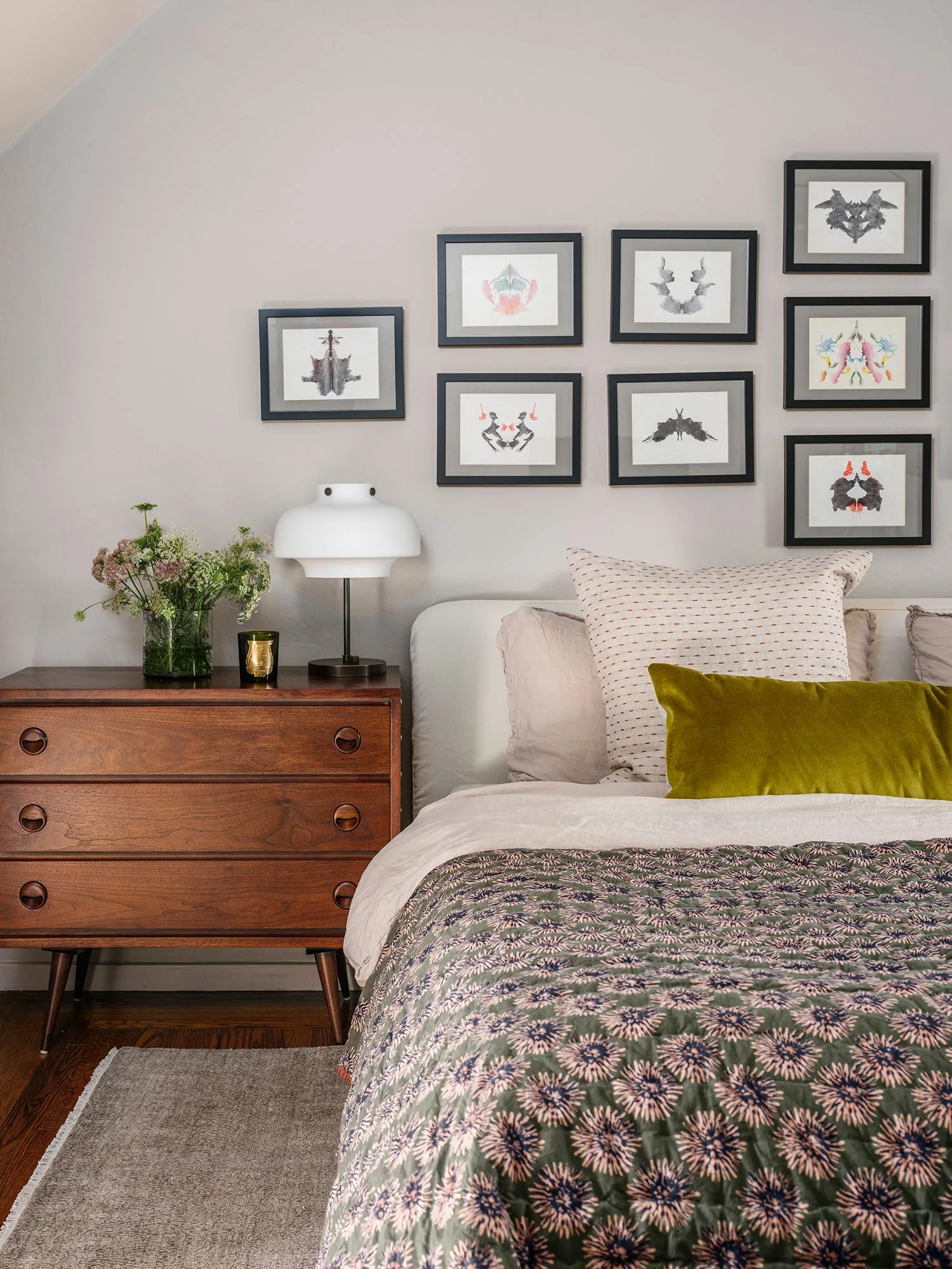 An eclectic bedroom featuring a mid-century wooden dresser next to a bed with patterned bedding, topped by a grid gallery wall of framed art.