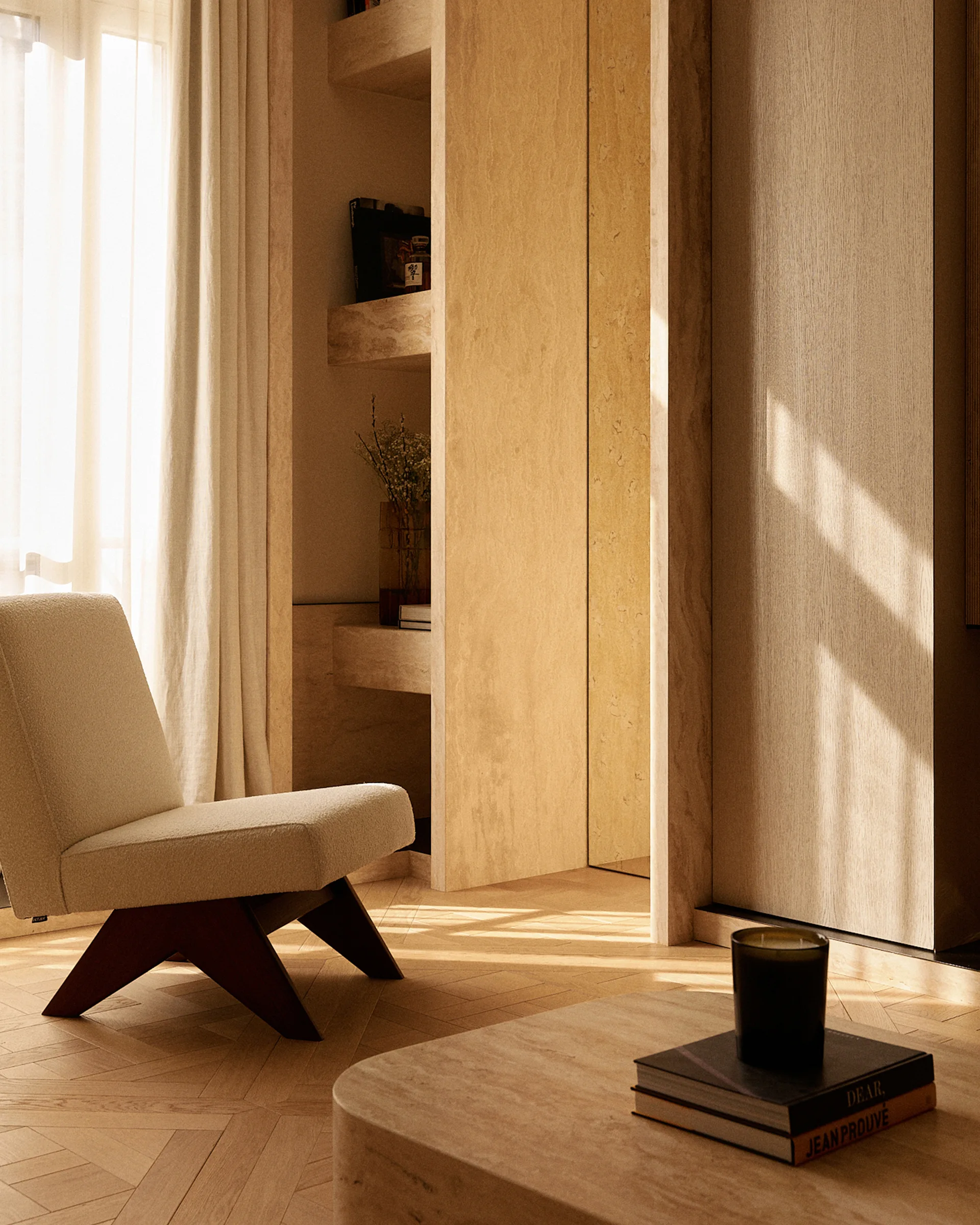 A sunlit corner features an upholstered armchair with dark wood legs, herringbone wood floors, and textured beige stone built-in shelving.