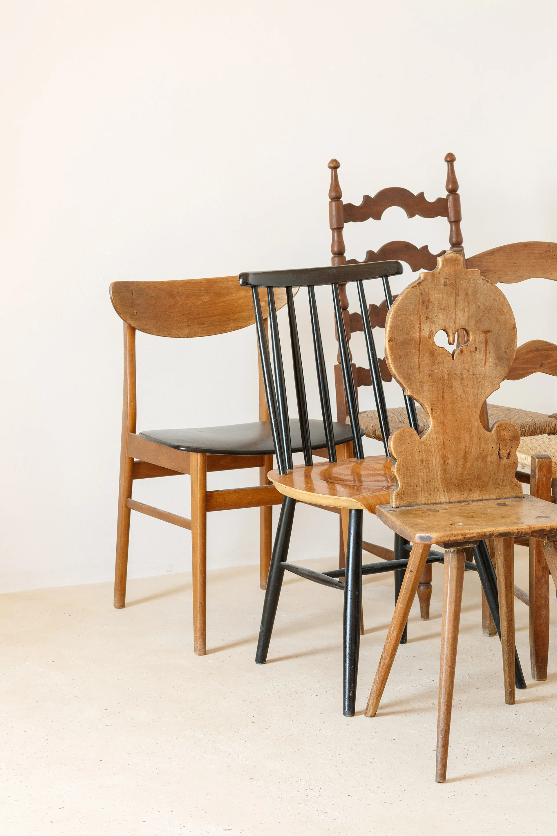 Five assorted vintage wooden dining chairs of varying styles and finishes are arranged against a plain white wall.