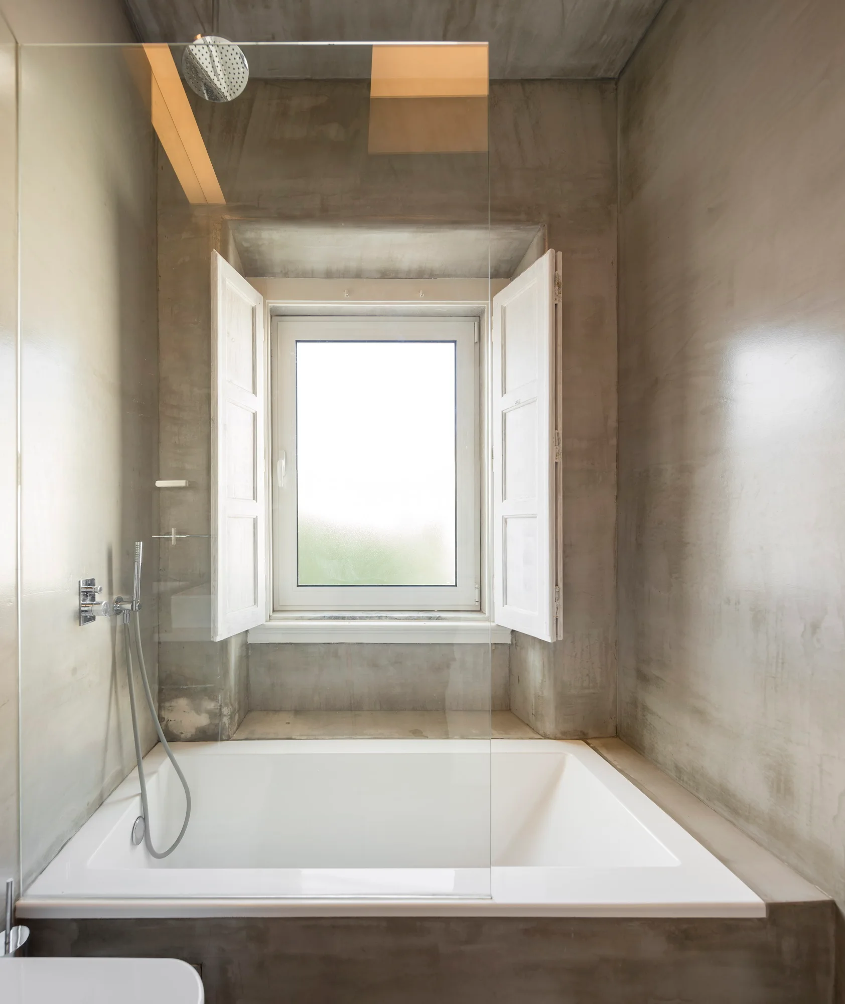 A modern concrete bathroom features a white built-in bathtub nestled below a window with open white wooden shutters and a glass shower panel.