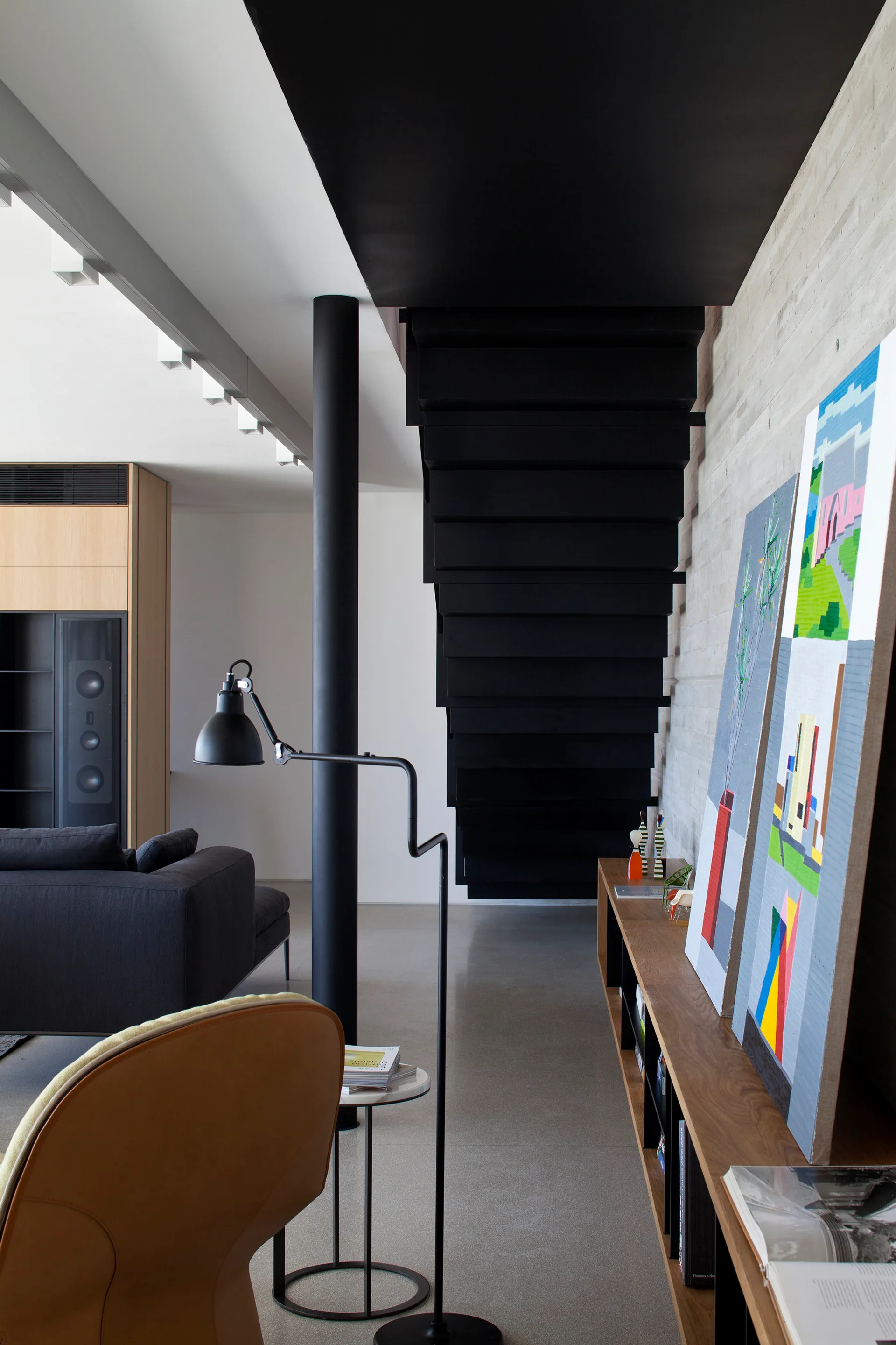 A black industrial staircase descends next to a textured concrete wall with a low wooden shelf holding leaning artwork in a modern living space.