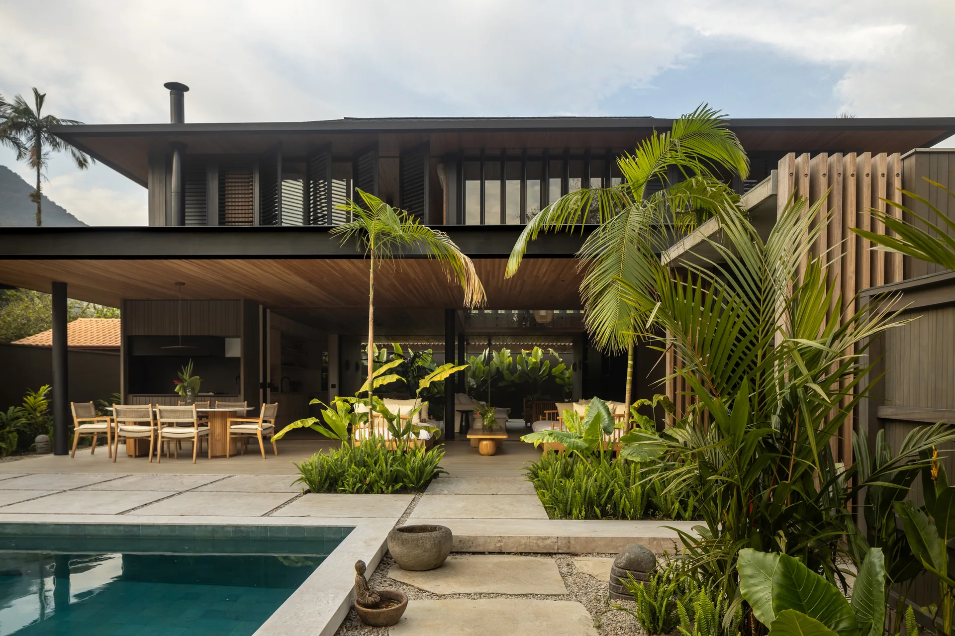 A modern two-story home features a covered wooden patio with a dining table and lounge seating next to a pool surrounded by tropical plants.