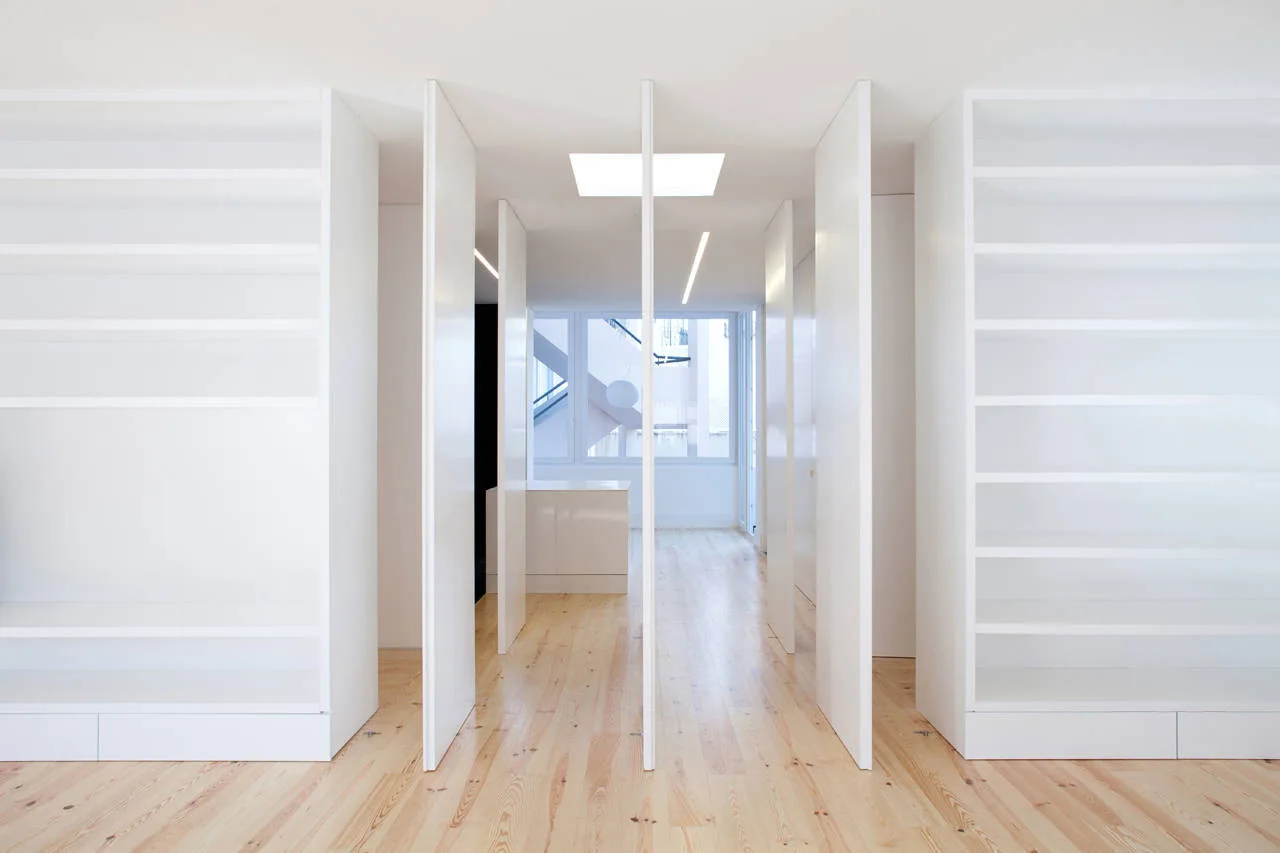 A minimalist white hallway features light wood floors, tall vertical pivoting doors, a square ceiling skylight, and large built-in shelving units.