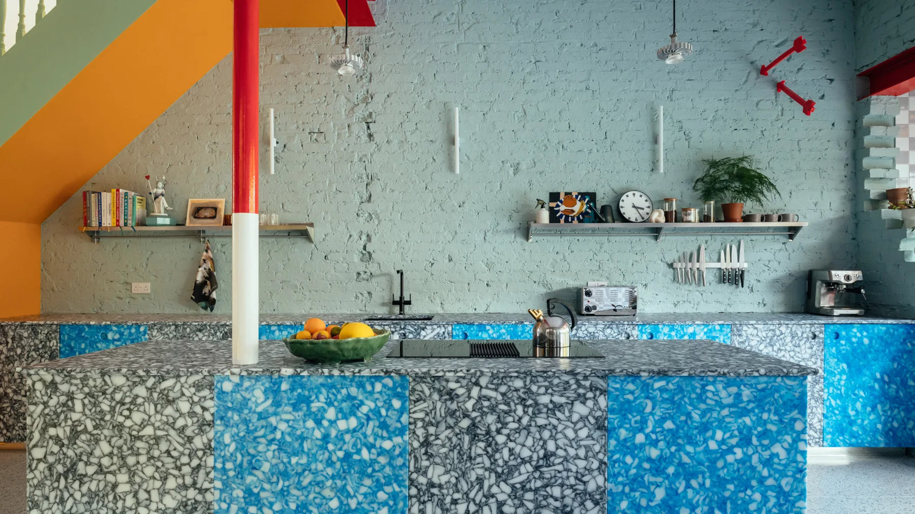 A contemporary kitchen features a blue terrazzo island and cabinets against a mint green painted brick wall with steel open shelves.