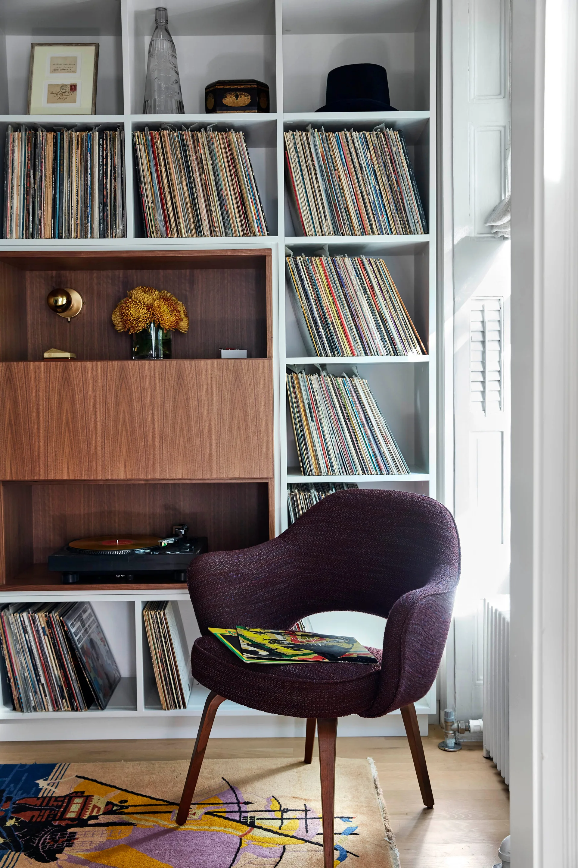 A white built-in bookcase filled with vinyl records features a wood-accented turntable cabinet next to a dark purple upholstered armchair.