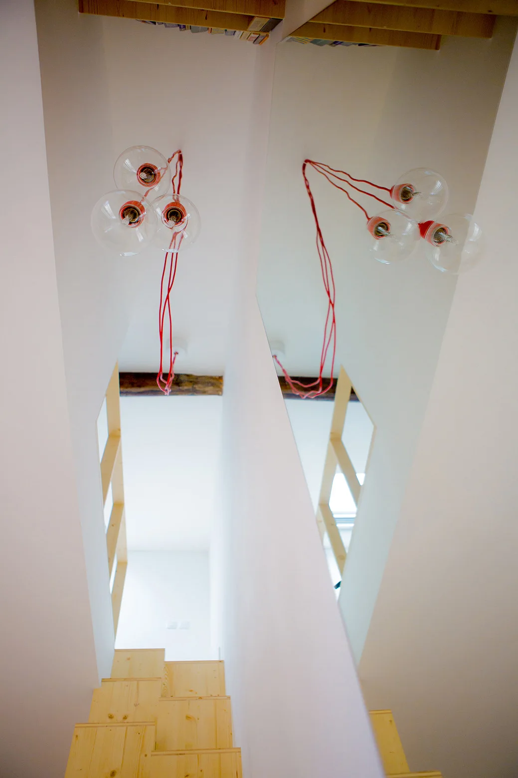 A modern wooden staircase with light-colored steps, featuring a large wall mirror and a pendant light with red cords hanging from the white ceiling.