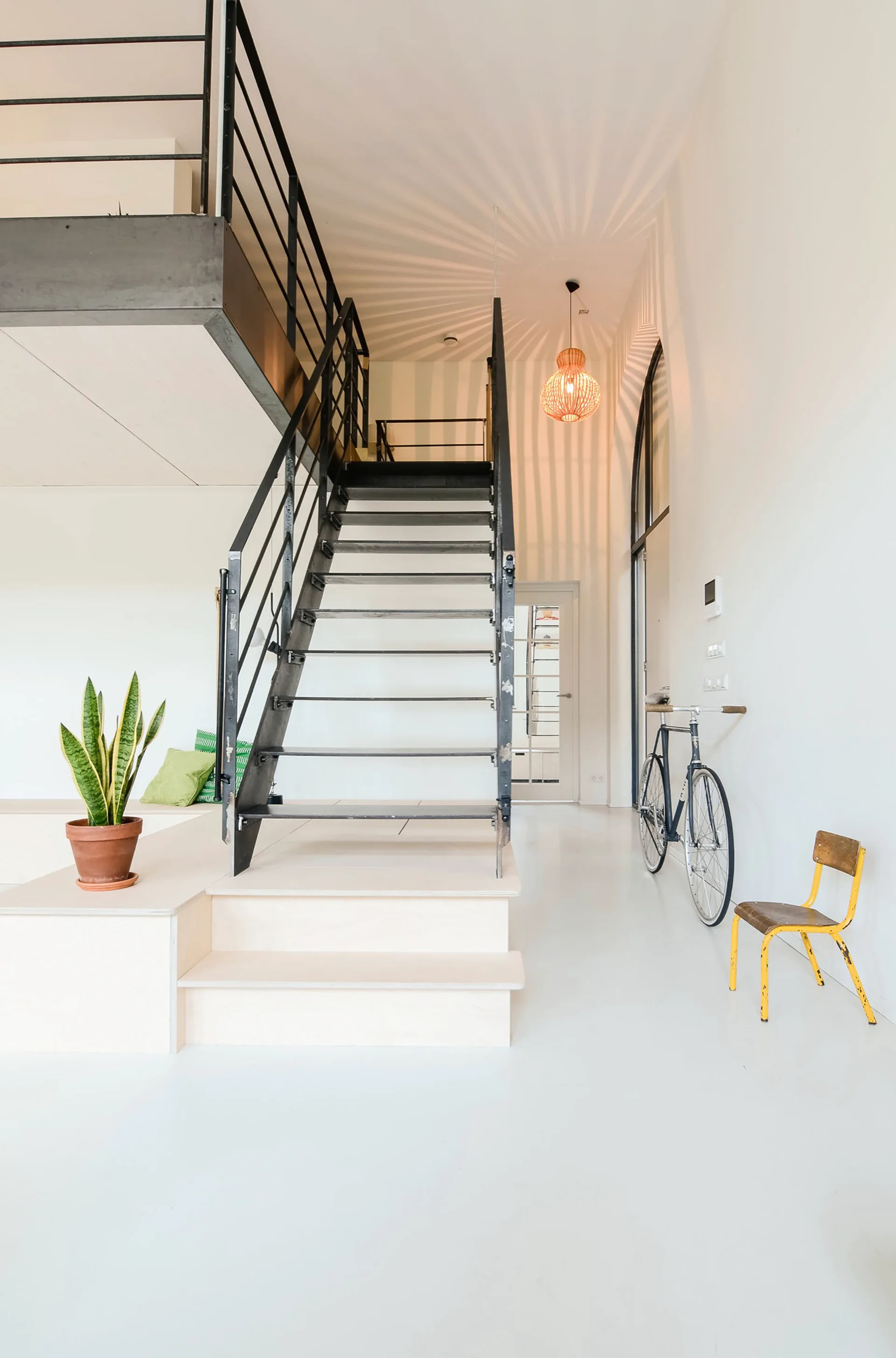 An industrial black metal staircase rises from a light wood platform in a minimalist white room with a bicycle and a small yellow chair.