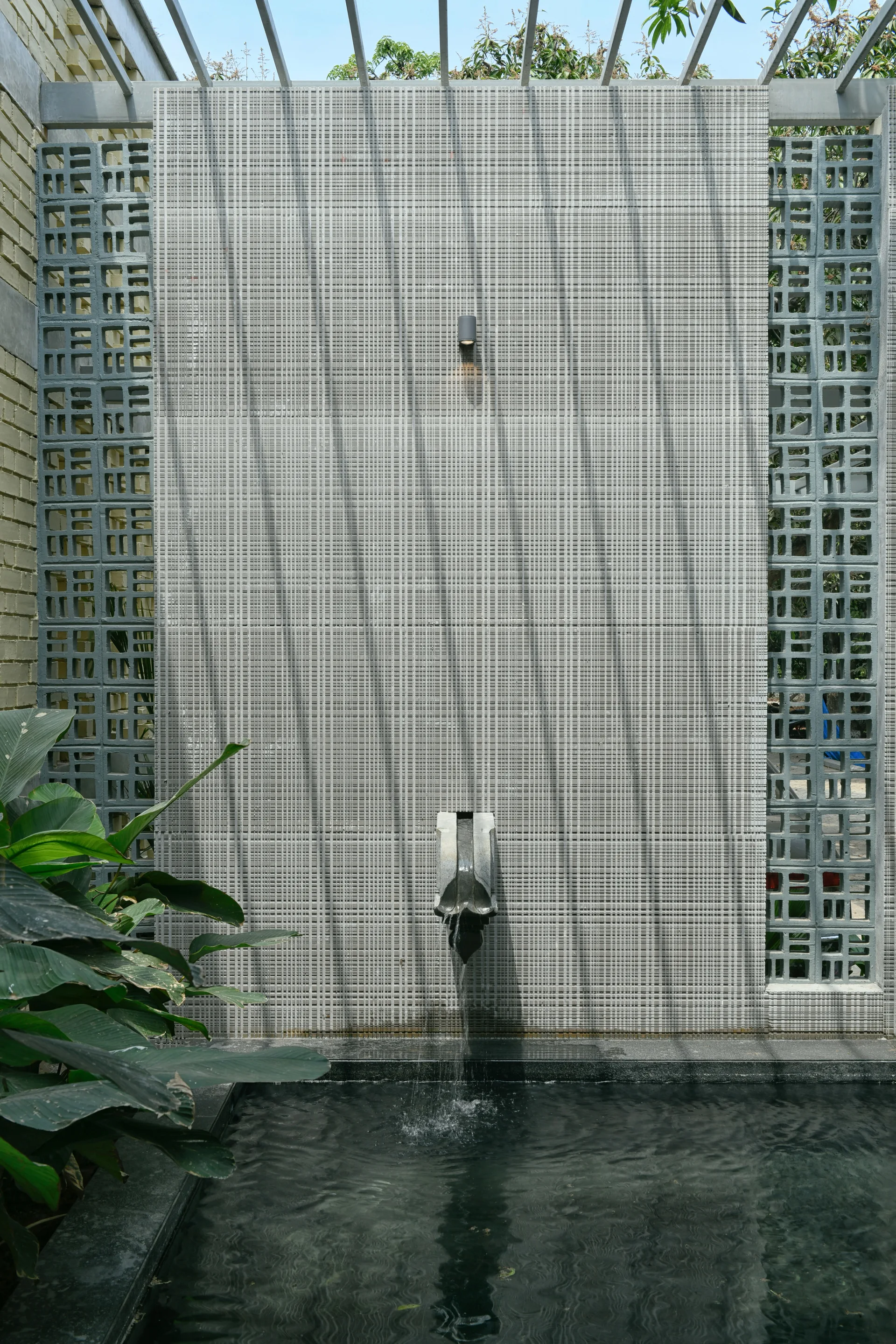 A modern outdoor water feature with a stone spout spilling into a pool, set against a grid-tiled wall and concrete breeze blocks.