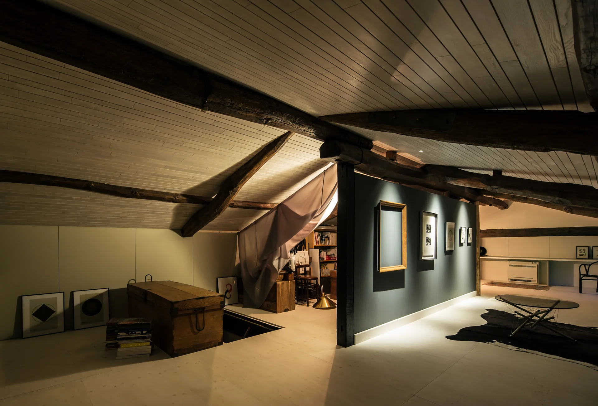 A dimly lit attic loft features a sloped white plank ceiling with dark wood beams, a central grey partition wall with framed art, and a wooden chest.