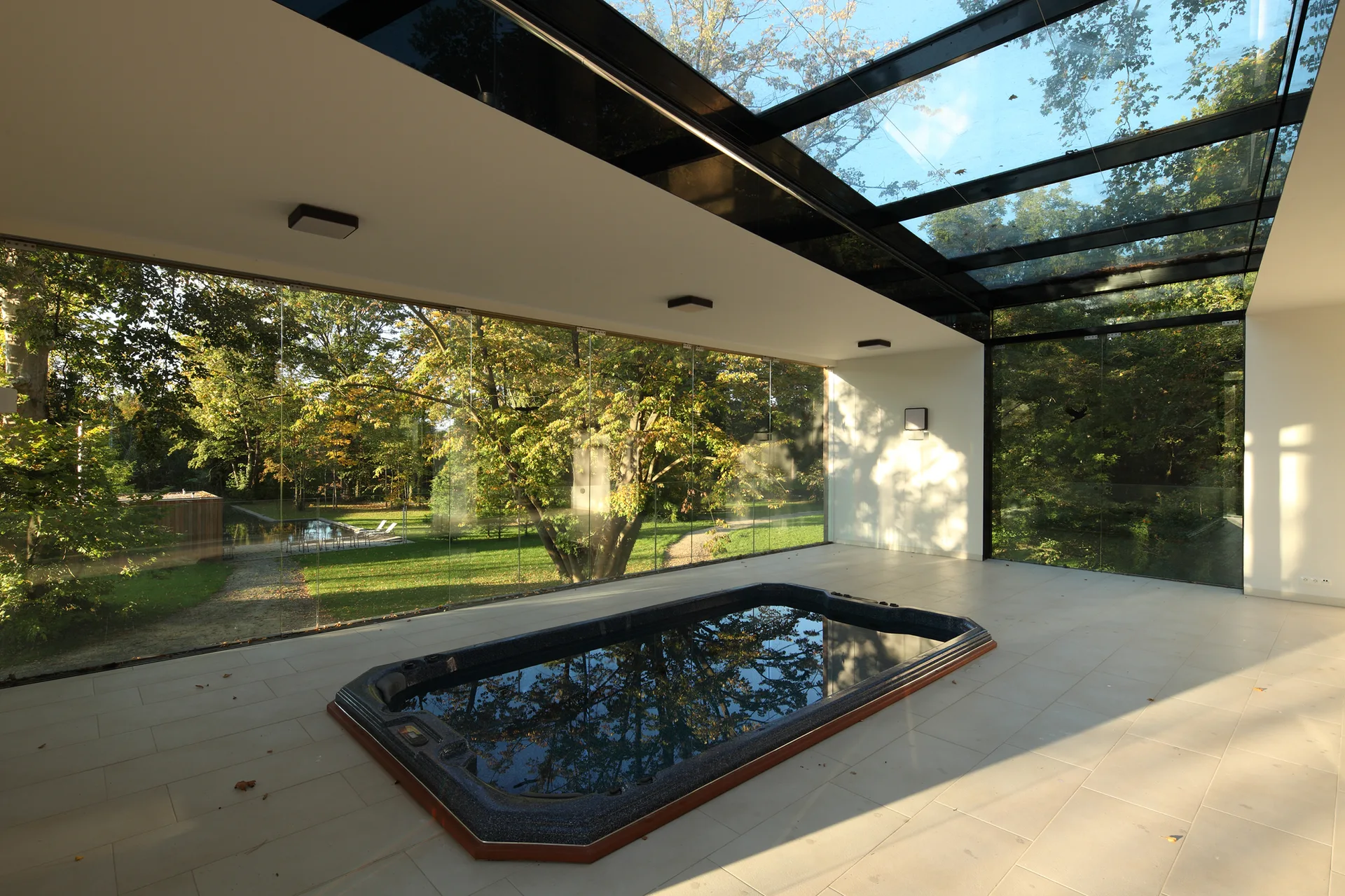 A modern indoor spa room with a recessed dark hot tub, large beige floor tiles, floor-to-ceiling glass walls, and a glass skylight ceiling.