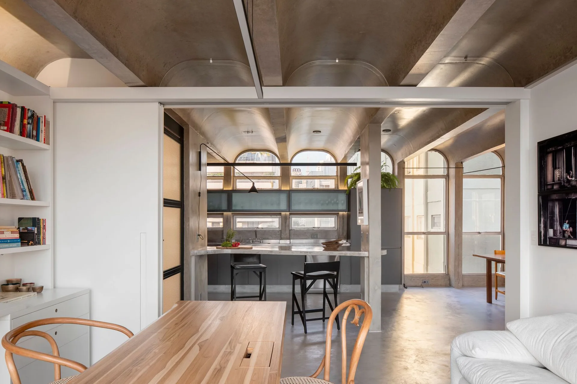 A modern open-plan interior features a concrete barrel-vaulted ceiling above a gray kitchen island with bar stools and a wooden dining table.