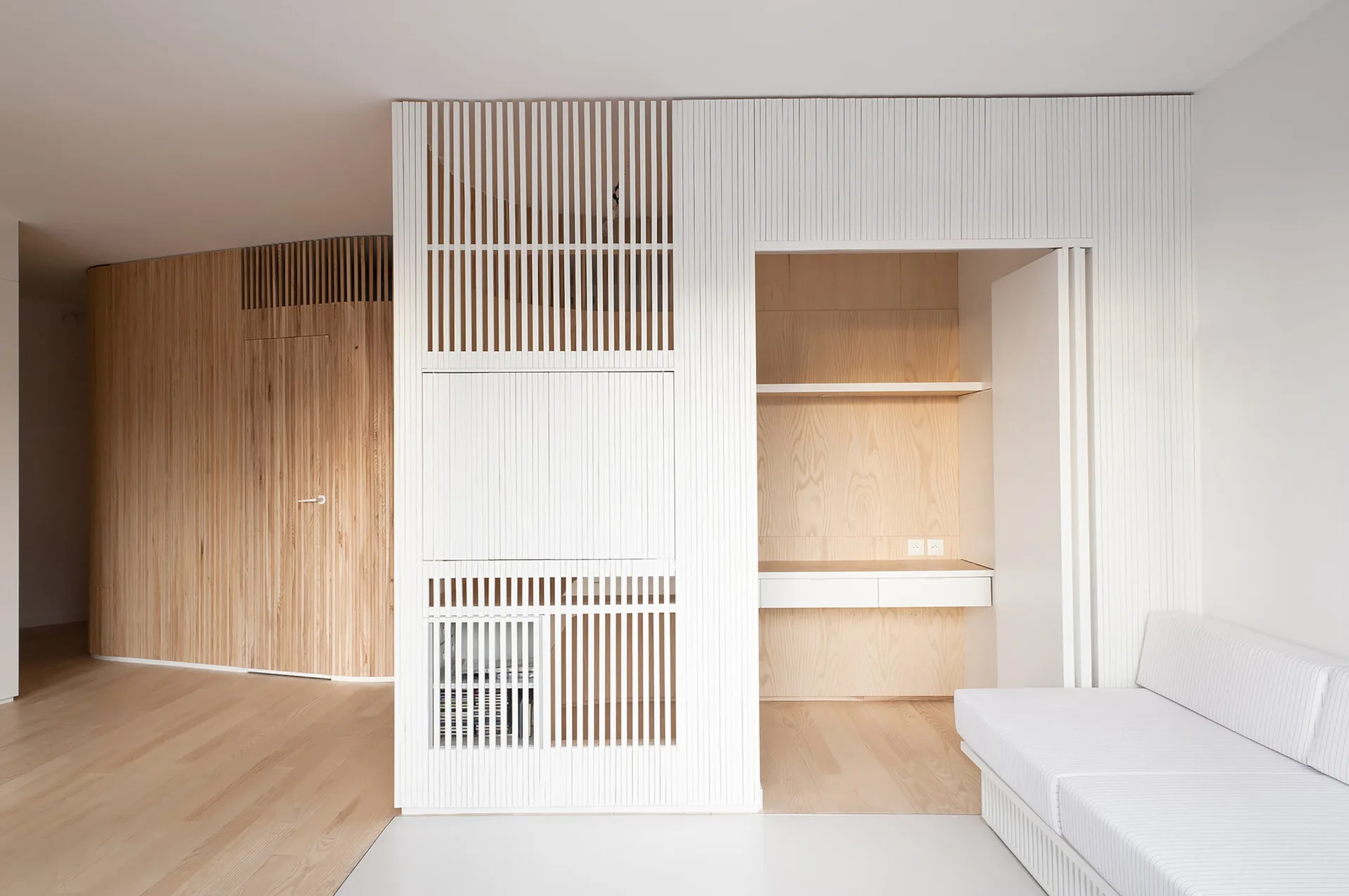 A minimalist room features a white slatted partition wall with a built-in wood desk nook next to a light wood floor and white sofa.
