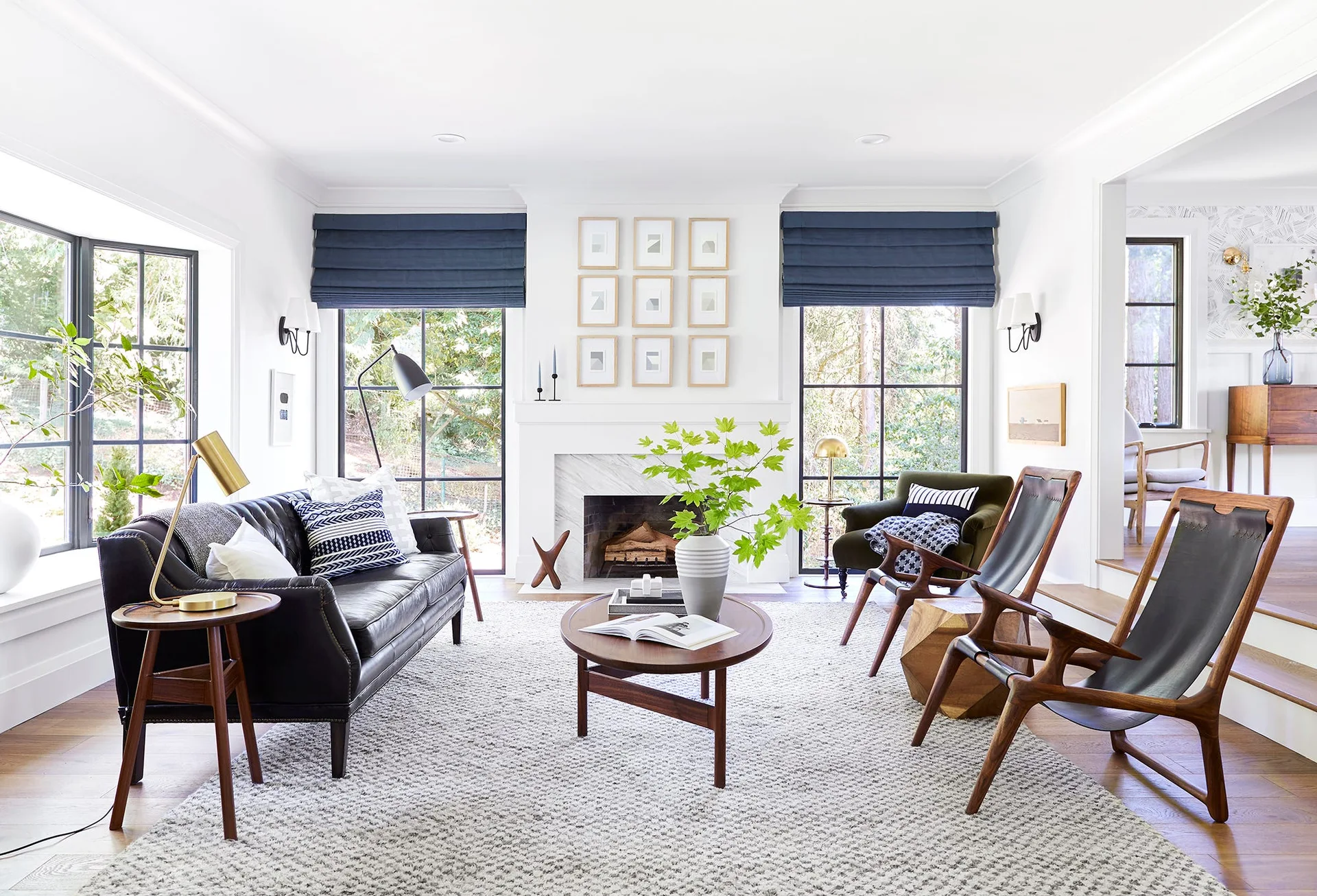 A white living room features a black leather sofa, wood sling chairs, and two large windows with navy blue roman shades flanking a fireplace.