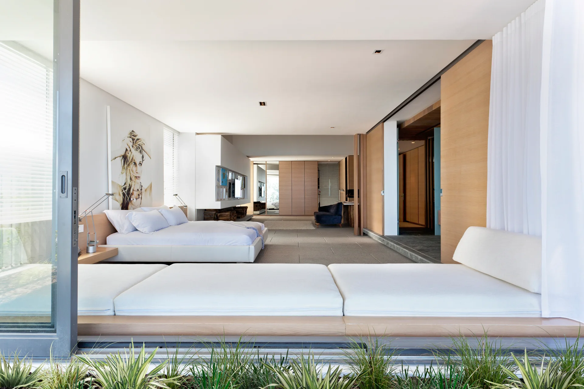 A minimalist modern bedroom features a low platform bed, integrated daybed seating by sliding glass doors, and light wood wall panels.