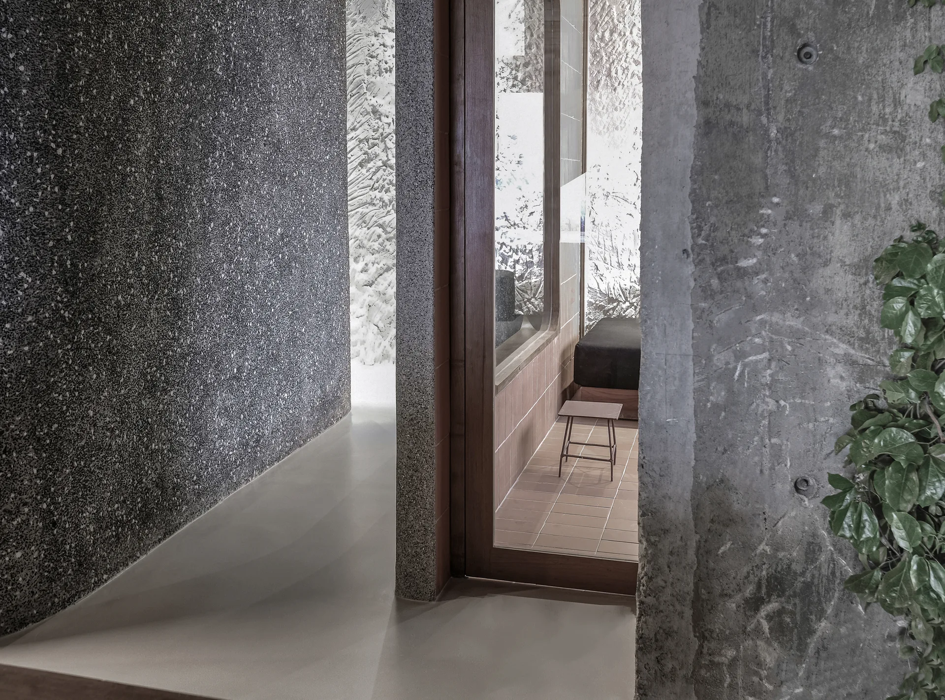 A minimalist interior passageway features a dark terrazzo wall, an exposed concrete partition, and a wood-framed glass door opening to a tiled room.