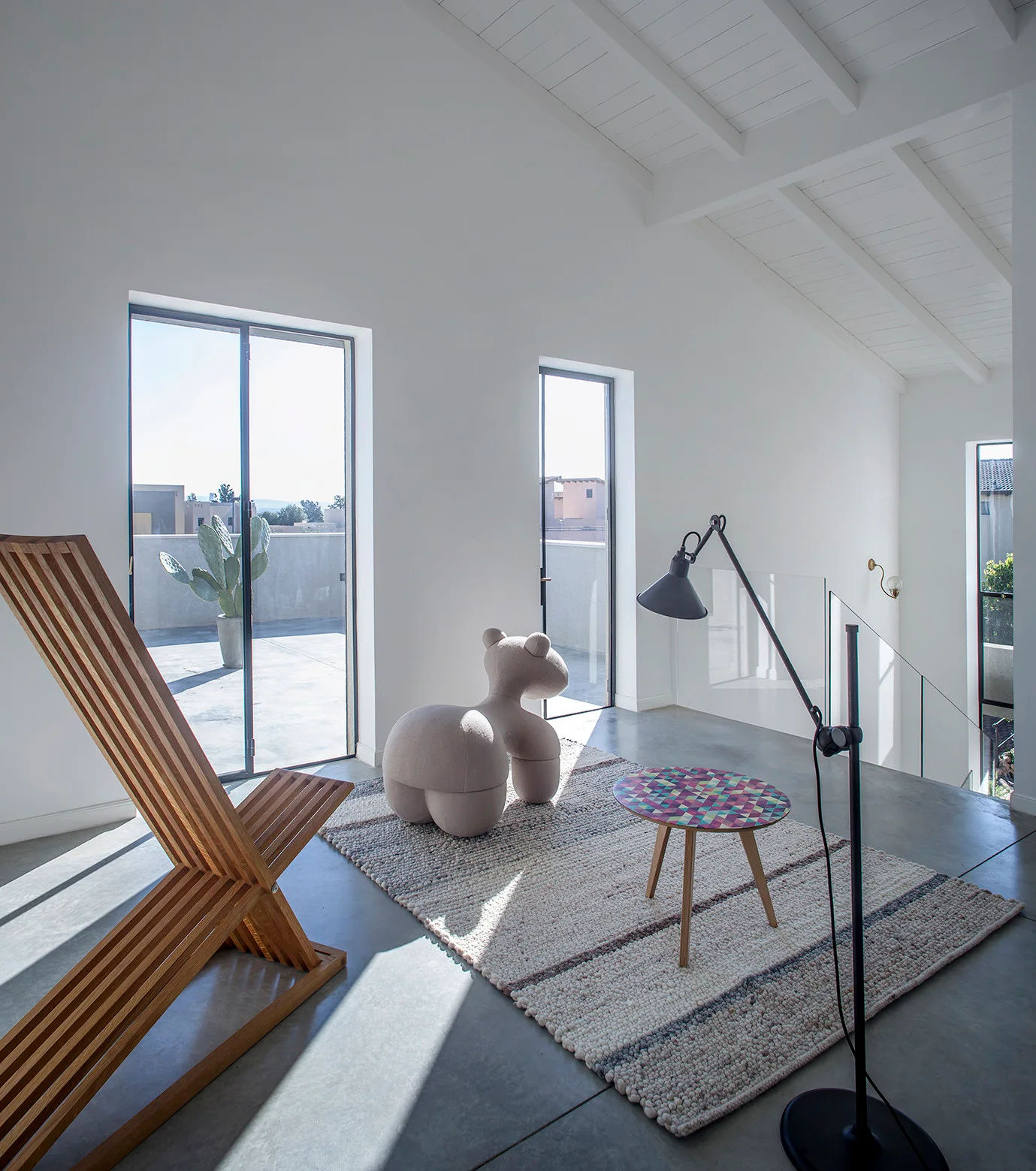 A modern loft living area features a wooden slatted chair, a beige animal-shaped lounge chair, a small mosaic table, and concrete floors.