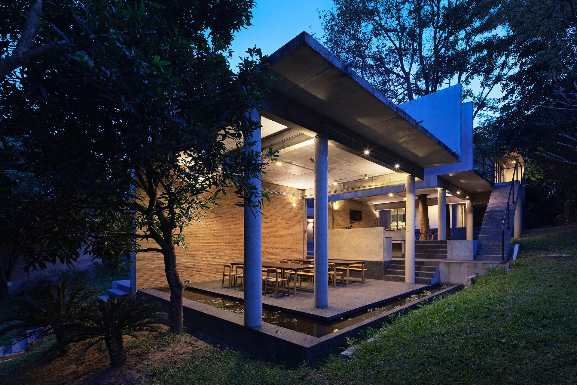 An open-air concrete patio features a long wooden dining table, a red brick accent wall, sturdy pillars, and a narrow reflecting pool.