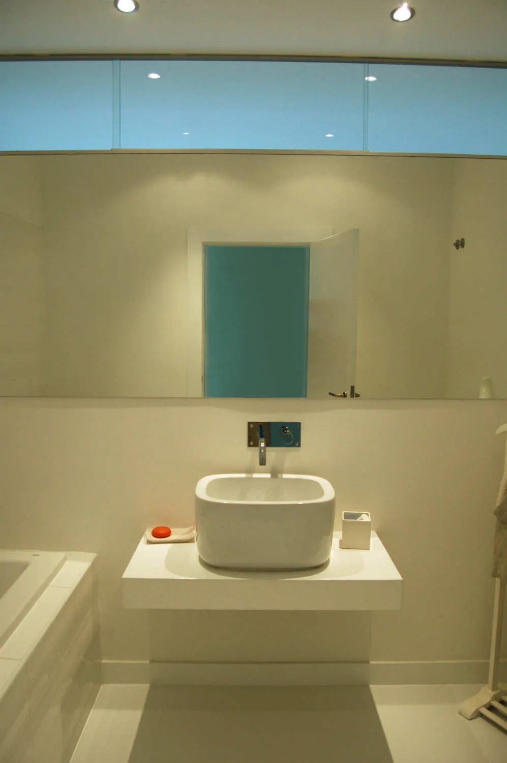 A minimalist white bathroom featuring a floating white vanity with a ceramic vessel sink, a large frameless mirror, and a wall-mounted chrome faucet.