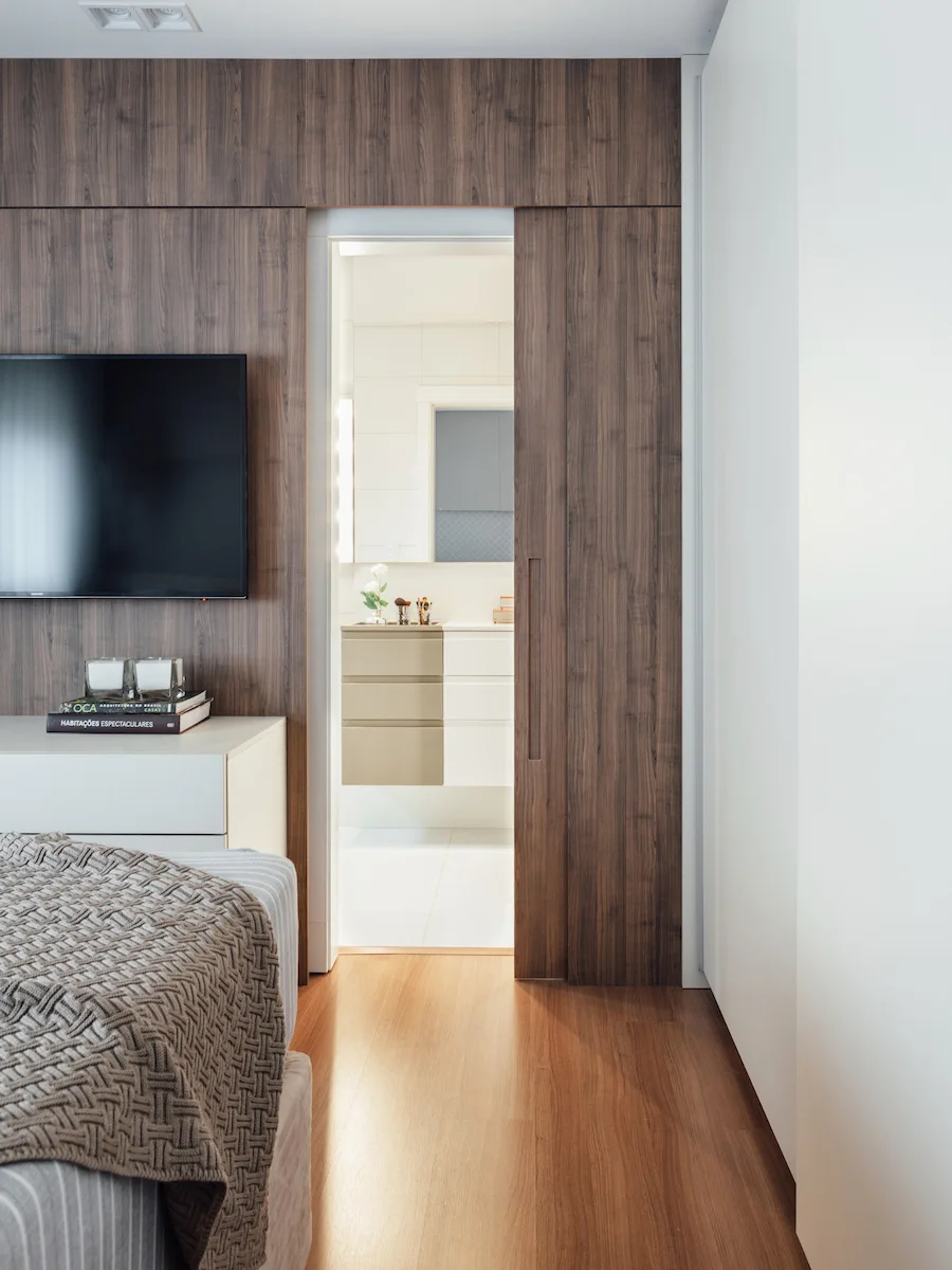 A modern bedroom features a warm wood-paneled wall with a matching sliding door that opens to reveal a bright, neutral-toned bathroom.