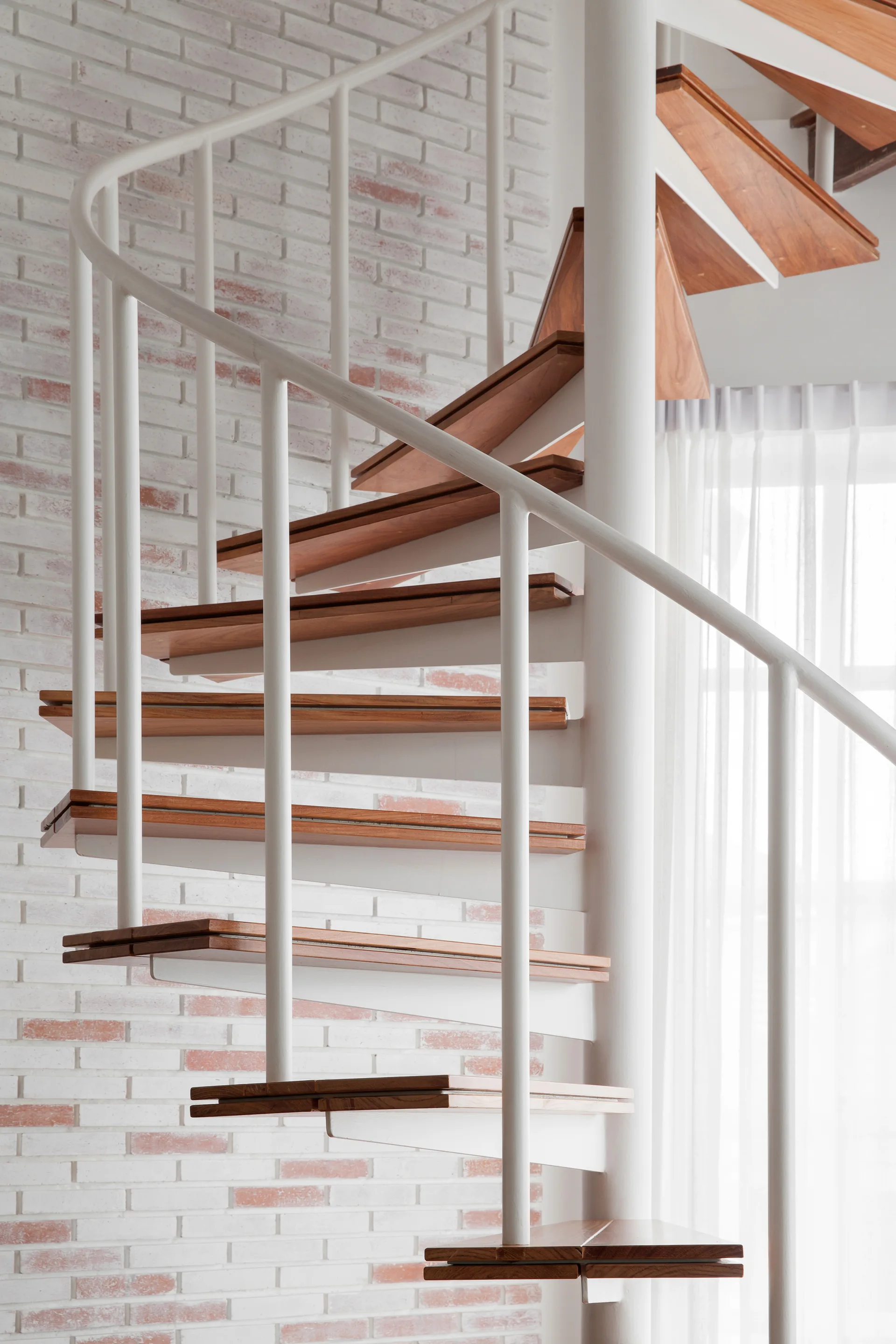 A close-up of a spiral staircase featuring rich wooden steps and a white metal railing set against a whitewashed brick wall.