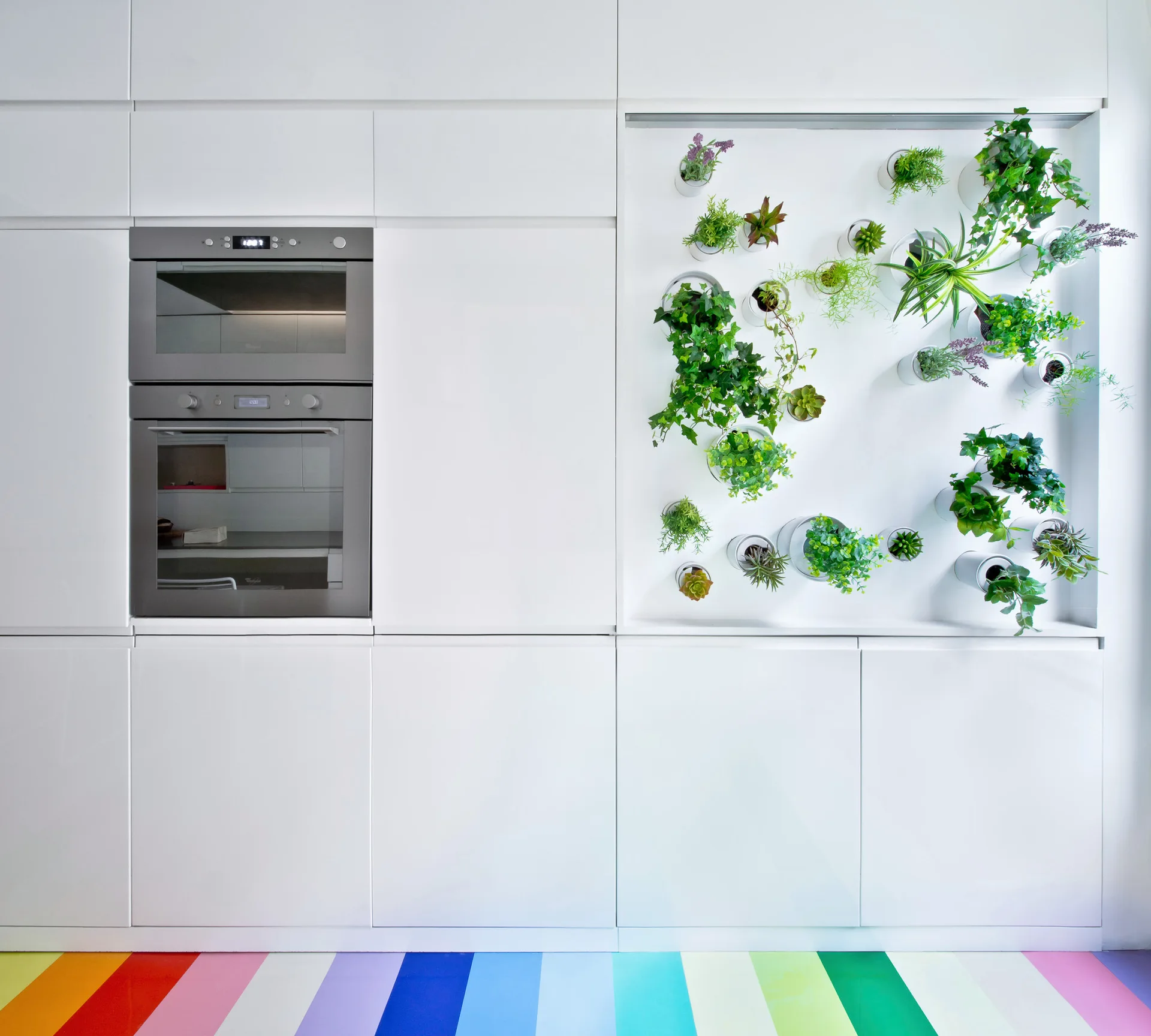 White minimalist kitchen cabinets feature an integrated double wall oven, a recessed vertical garden with small potted plants, and a rainbow striped floor.